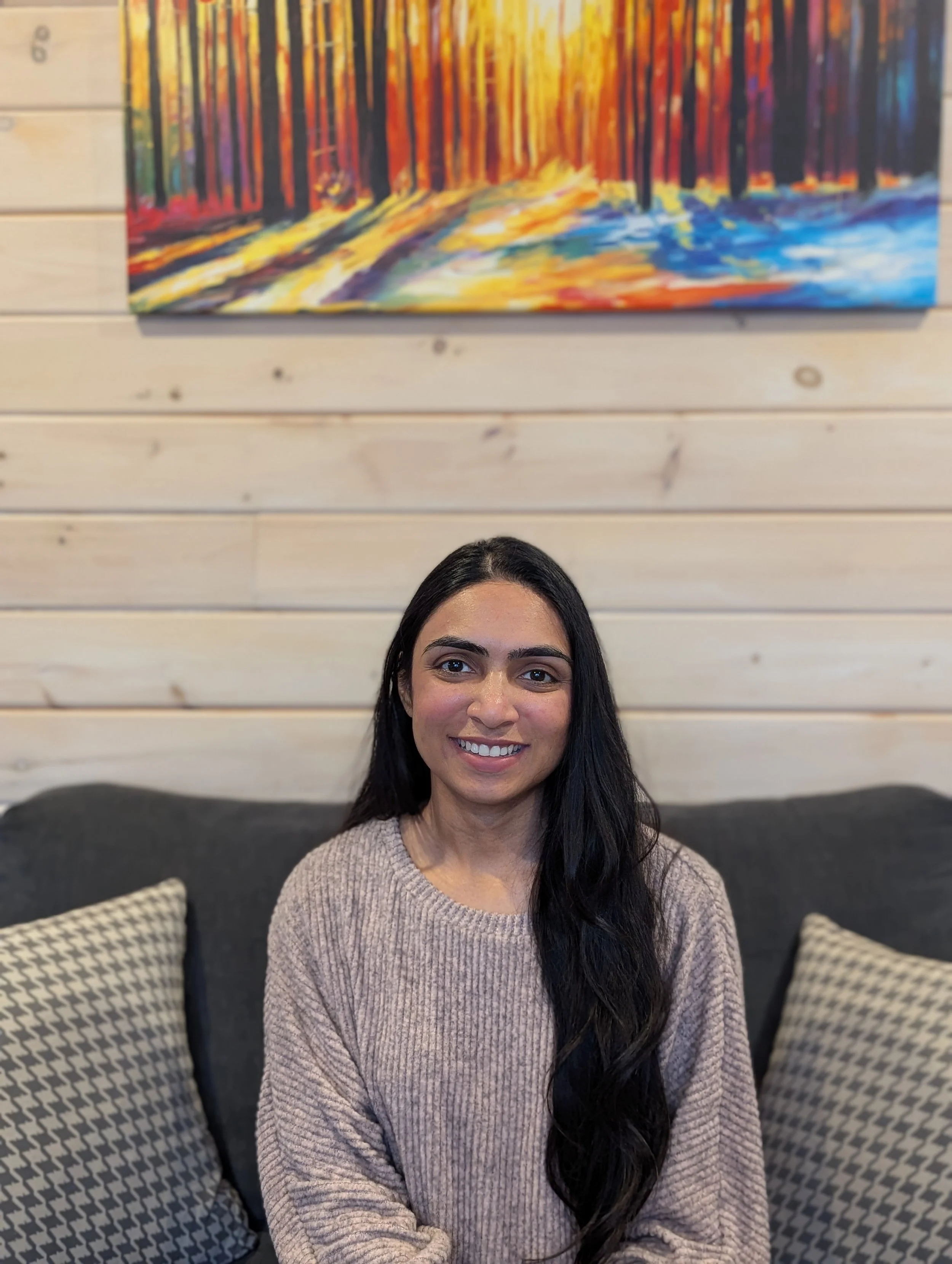 A woman with long dark hair and a beige sweater, sitting on a dark gray couch with patterned pillows, smiling at the camera in front of a light wood paneled wall with a colorful abstract forest painting.