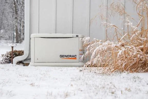 A Generac backup generator outdoors in snowy weather, next to a white fence and dried plants.