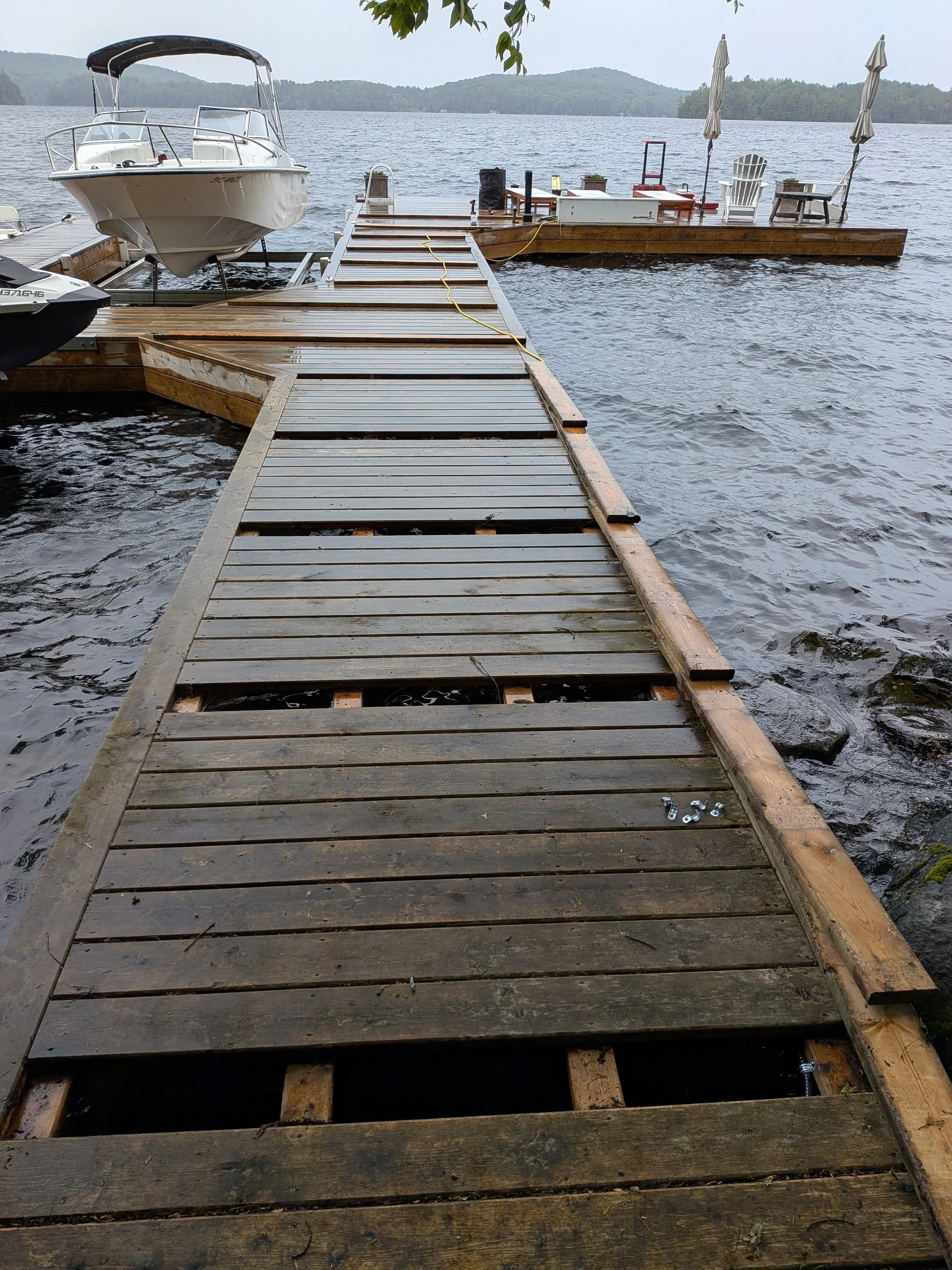 A wooden dock extending into a lake with a boat on the left side, and two Adirondack chairs with umbrellas, along with other outdoor furniture on the right side, during daytime with overcast skies and distant green hills.