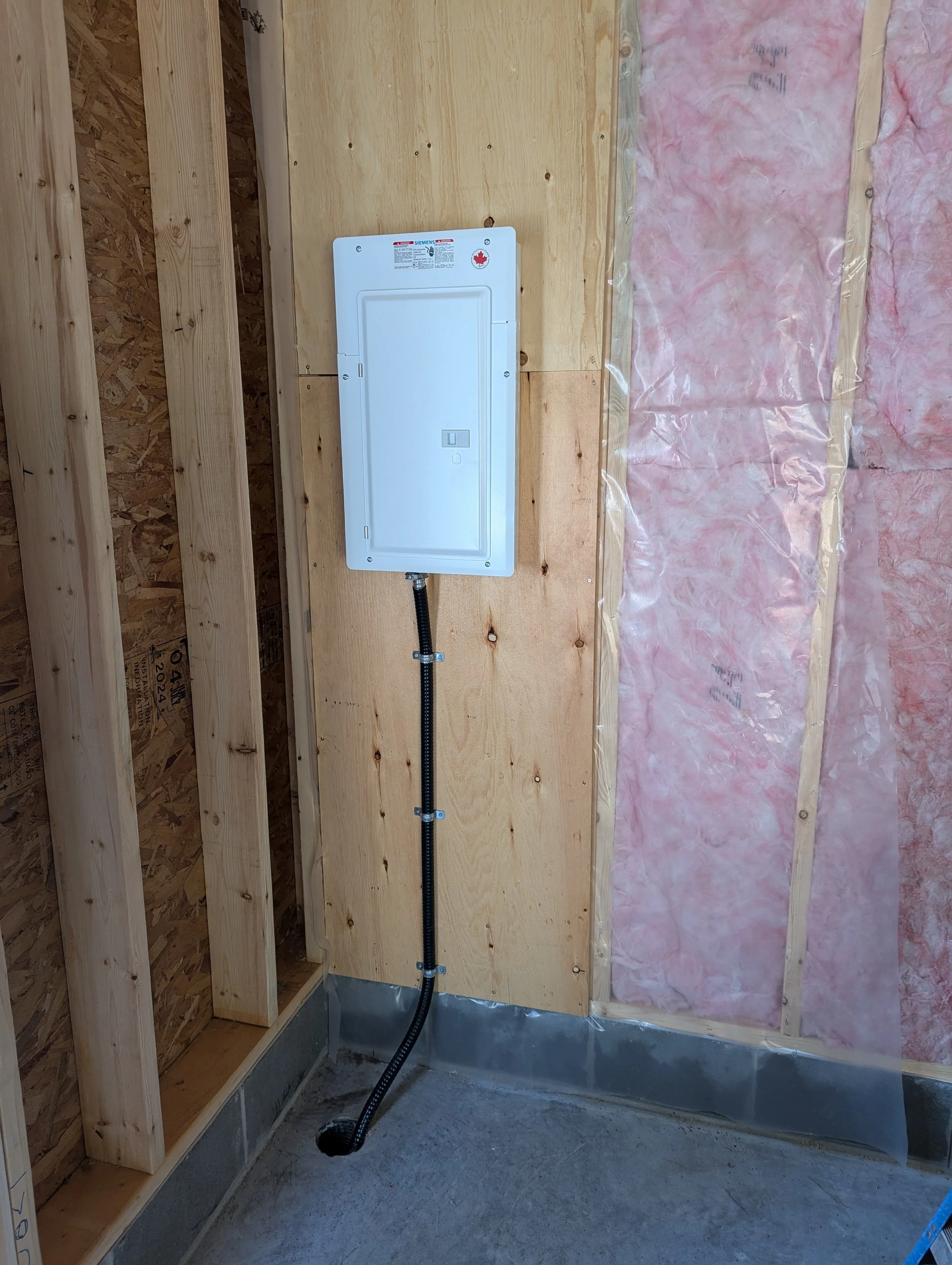 Electrical panel with a black conduit pipe below it installed on unfinished wooden wall with pink insulation on one side.