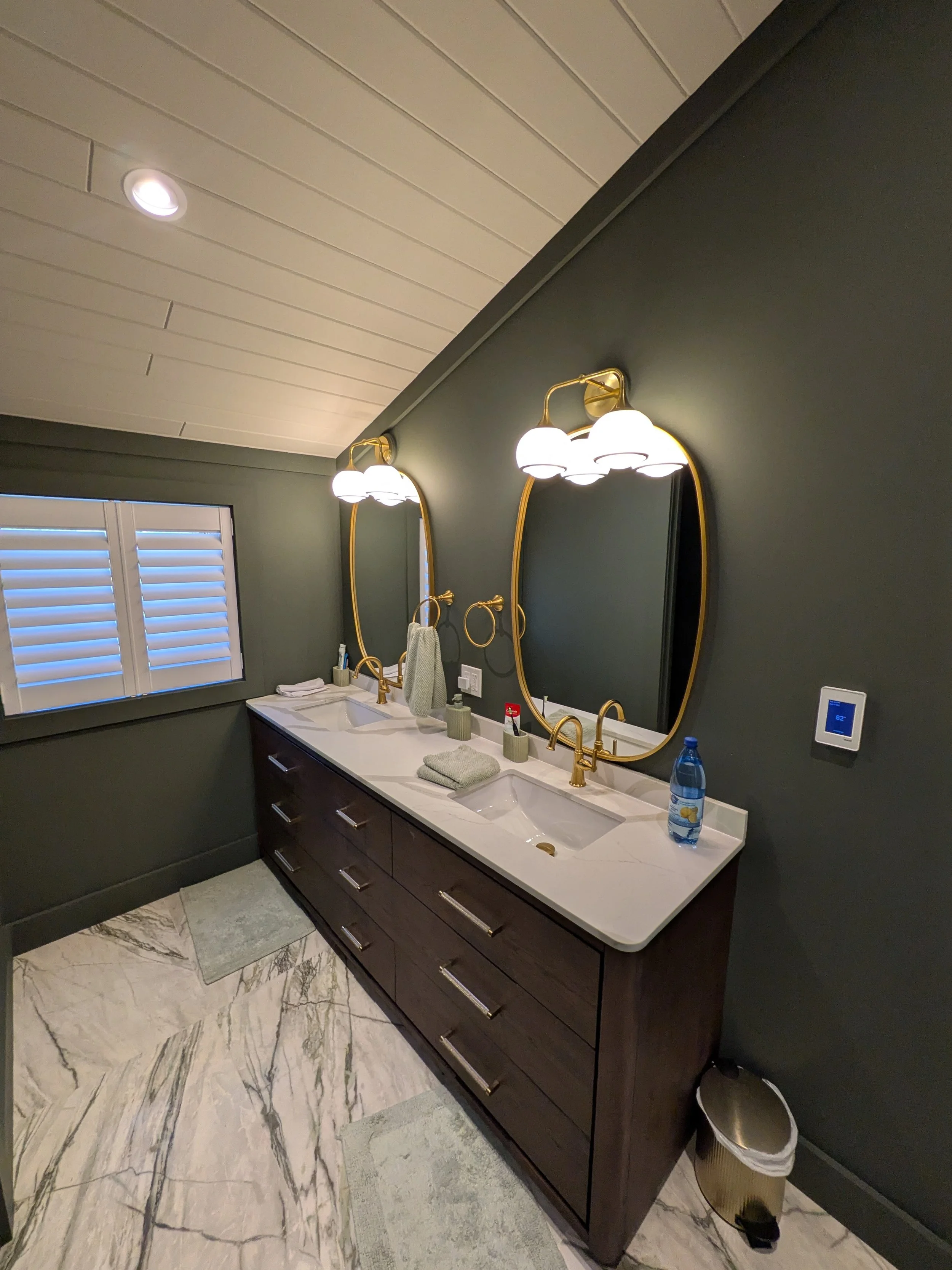 Modern bathroom featuring a double vanity with gold fixtures and matching oval mirrors, white marble countertops, green walls, window with plantation shutters, two pieces of gray towels, a bottle of water, and a trash bin.