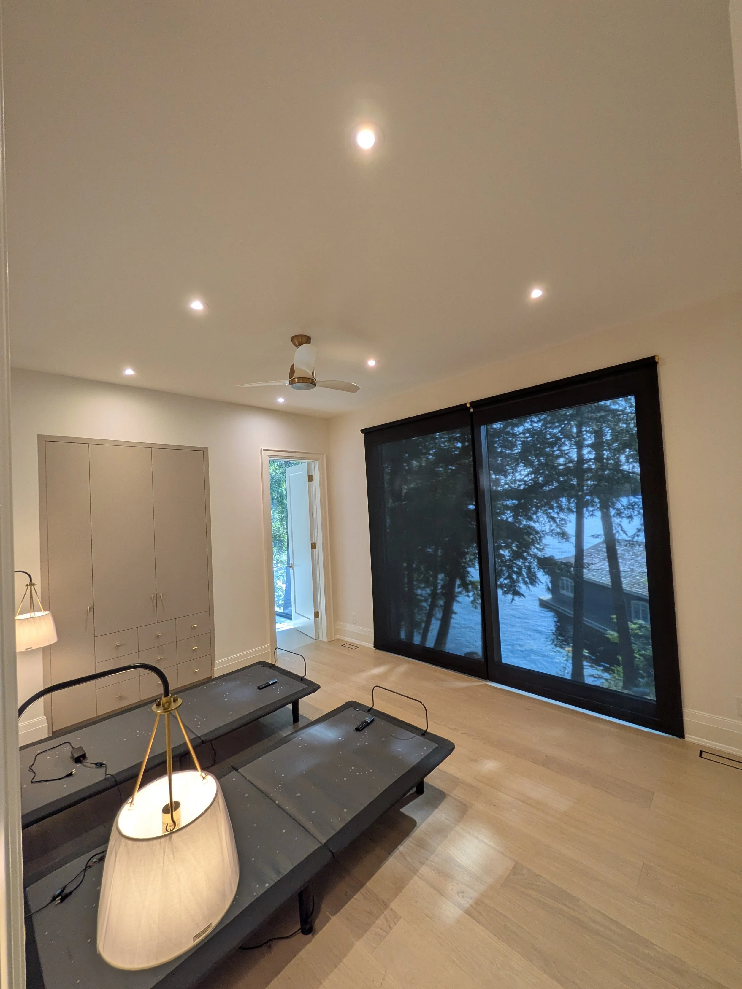 Empty living room with large window looking out to trees and lake, light wood flooring, beige walls, built-in beige cabinets, ceiling fan, and two black platform beds with scattered debris, lamp, and remote controls.