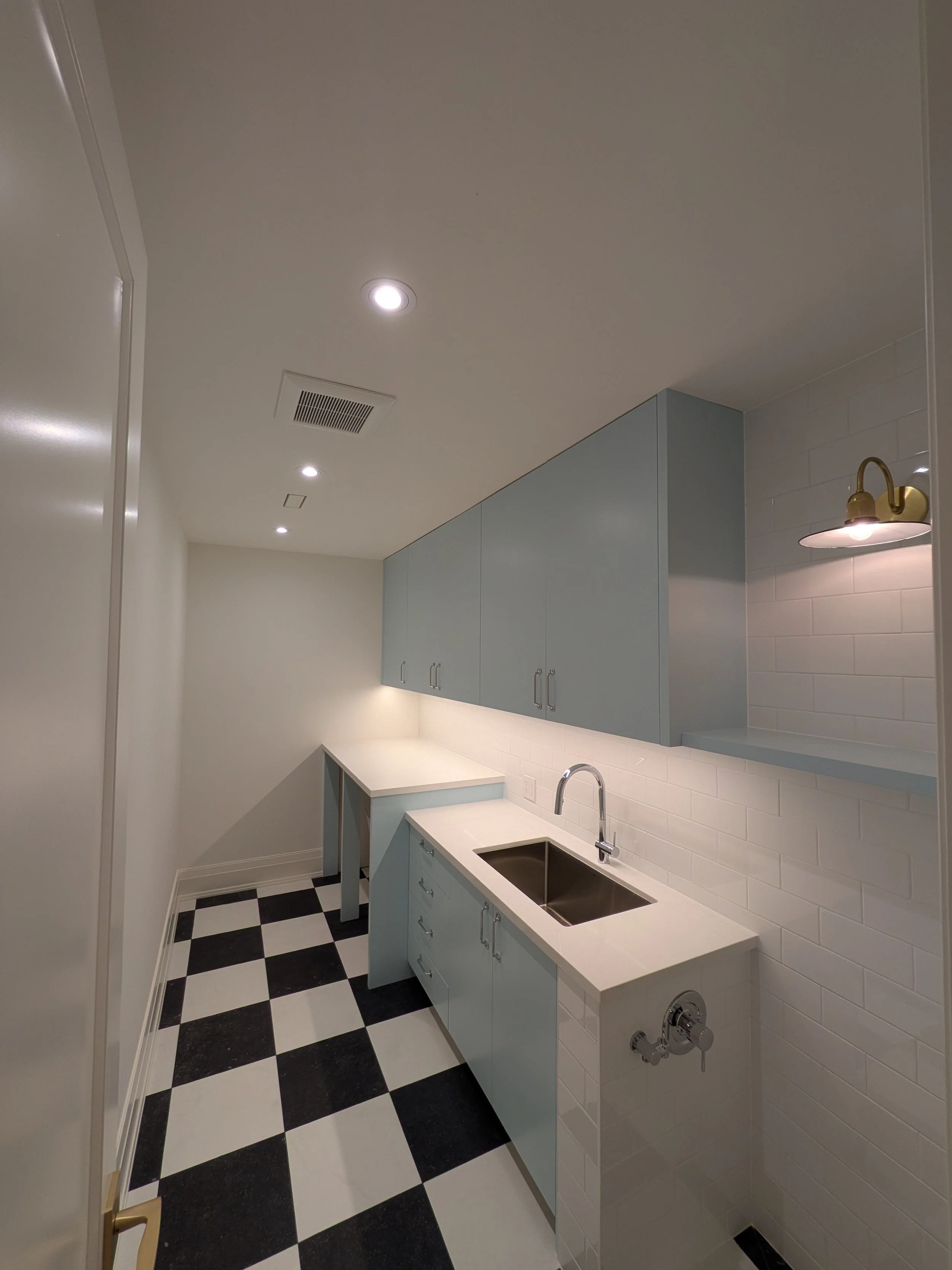 A compact kitchen with light blue cabinets, white countertops, black and white tile flooring, recessed ceiling lights, a small wall-mounted light fixture, and a stainless steel sink.
