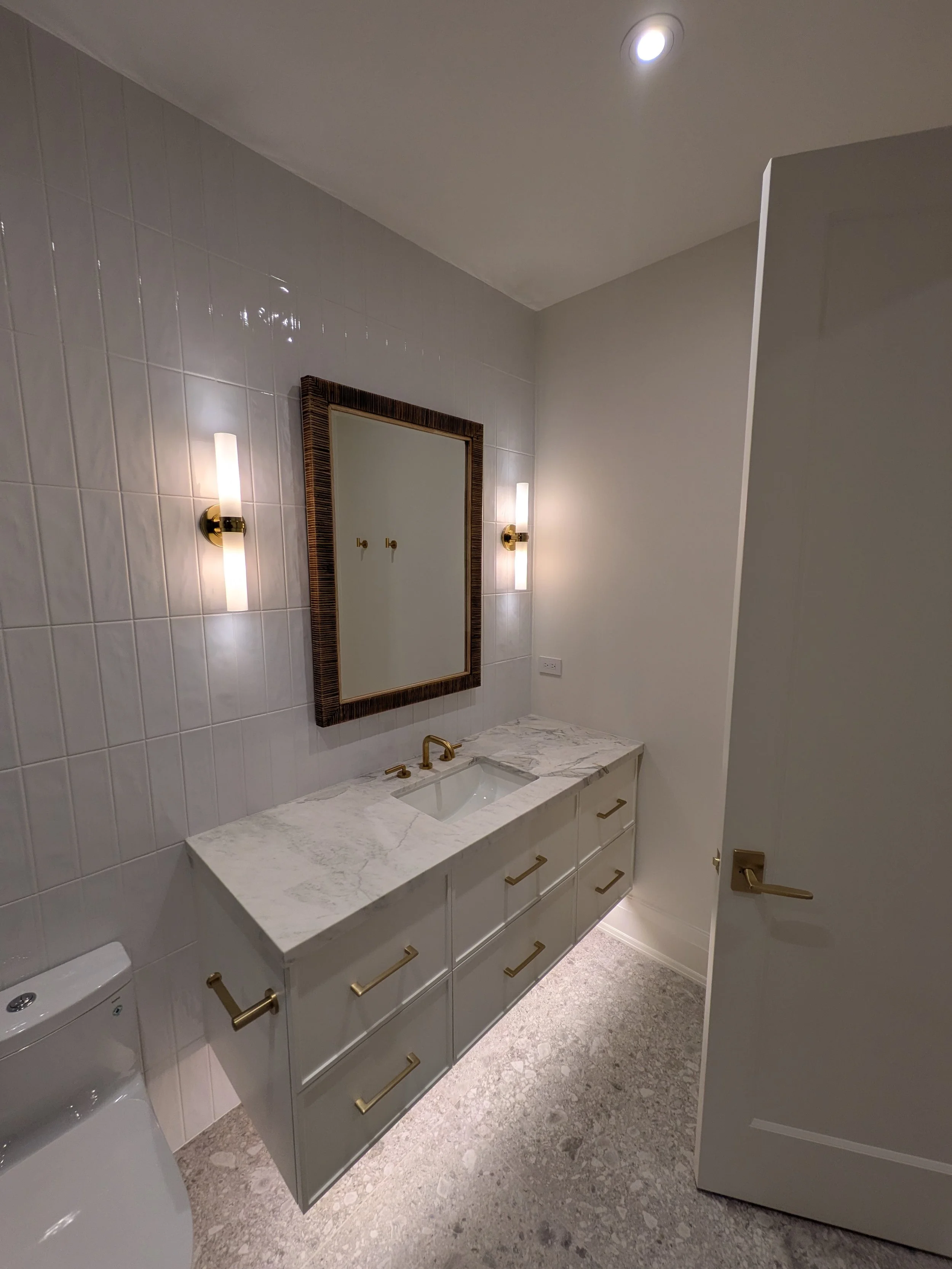 Modern bathroom with white vanity, marble countertop, gold fixtures, wall-mounted mirror, and wall sconces.