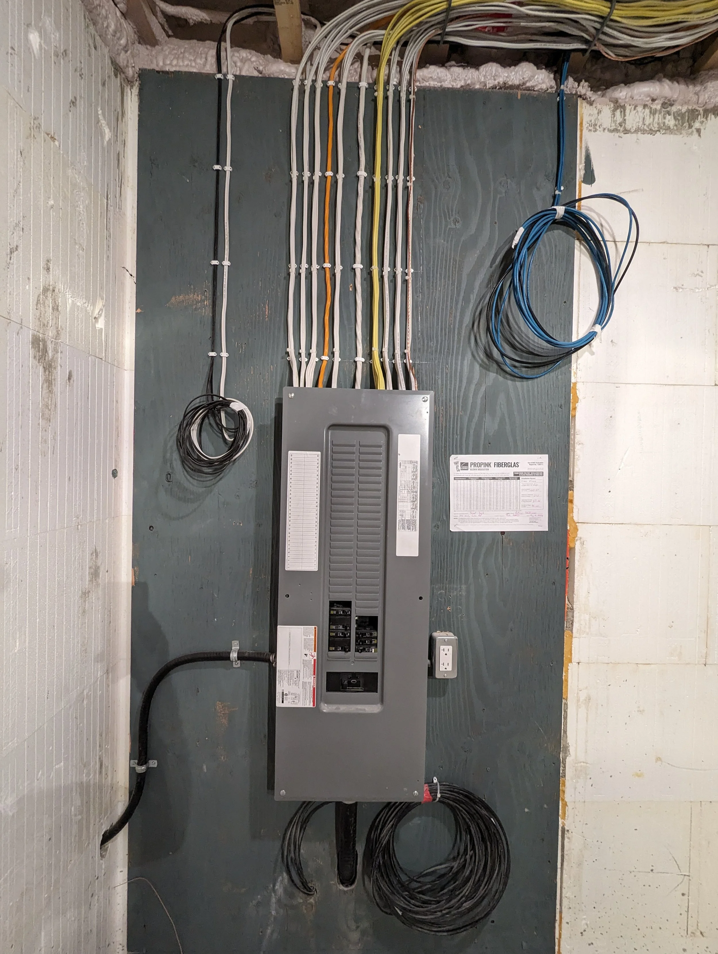 Electrical service panel with various colored wires organized and connected, mounted on a wall with electrical codes and labels.
