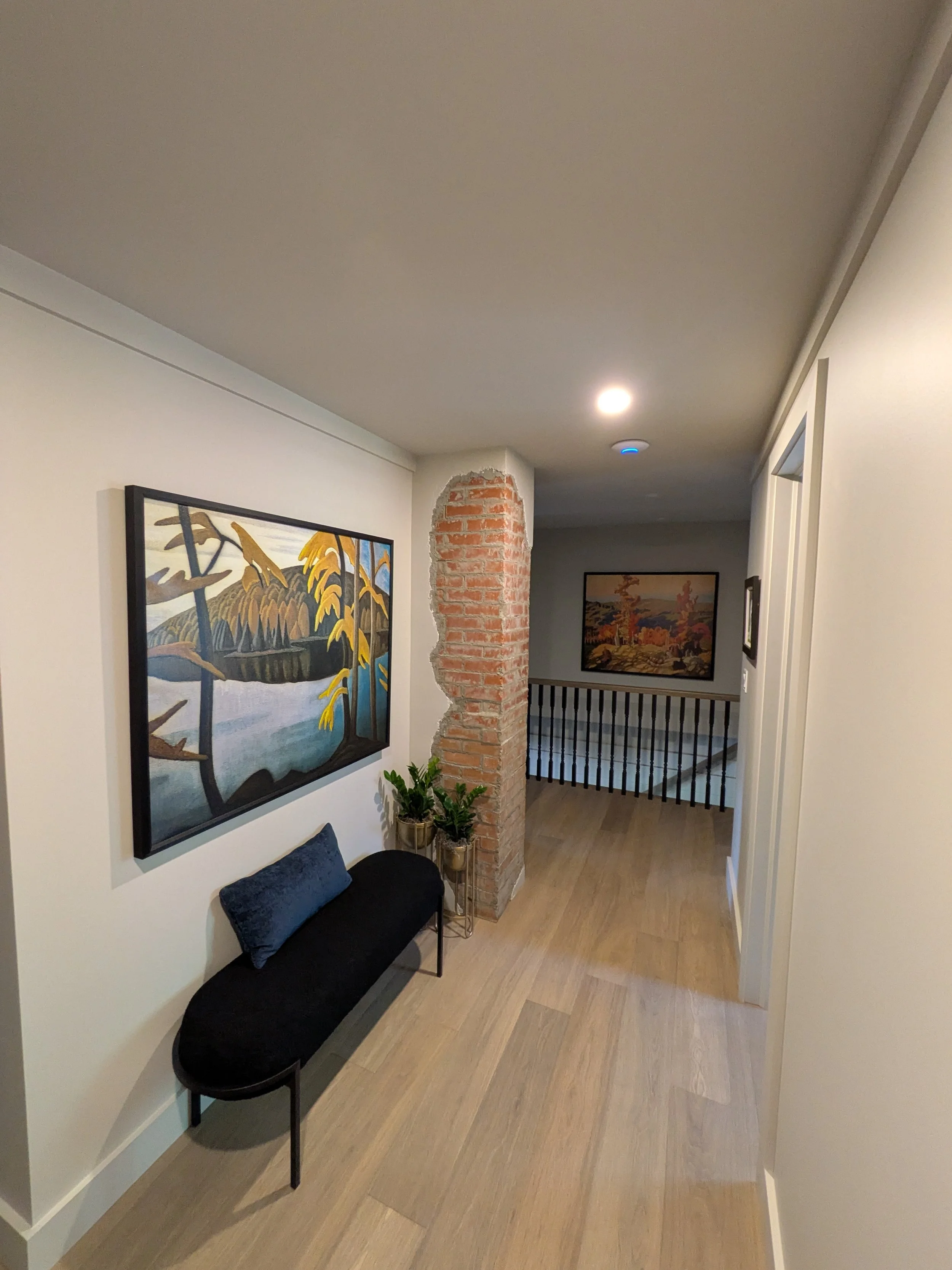 A hallway with a painting of a landscape on the wall, a small black bench with a blue cushion, two potted plants on gold stands, a partially visible brick column, and another painting at the end of the hall.