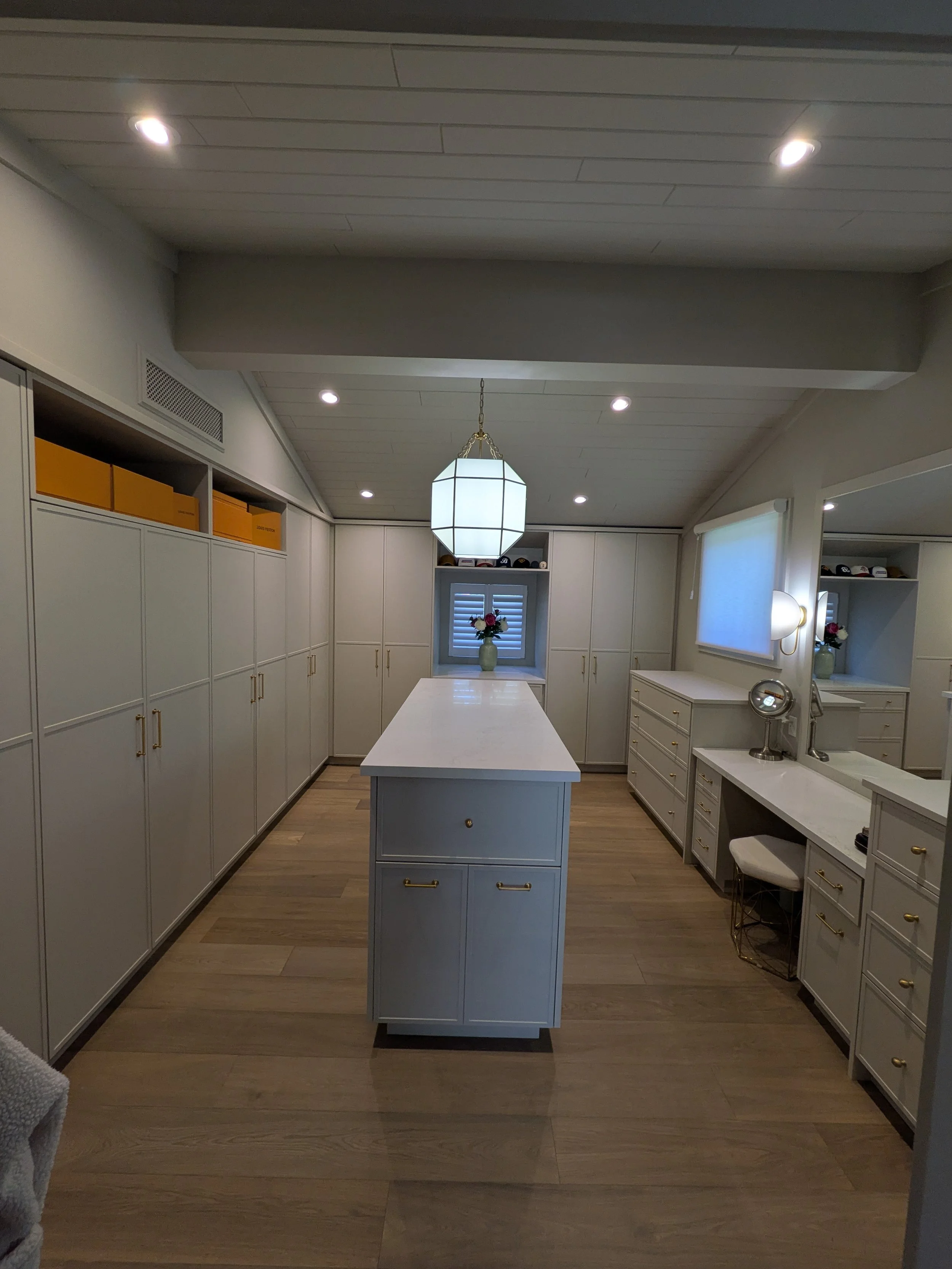 A spacious, well-lit walk-in closet with white cabinetry, gold handles, and a central island with drawers. There are built-in cabinets along the walls, a window with shutters at the far end, and decorative items including a vase with flowers. The roo