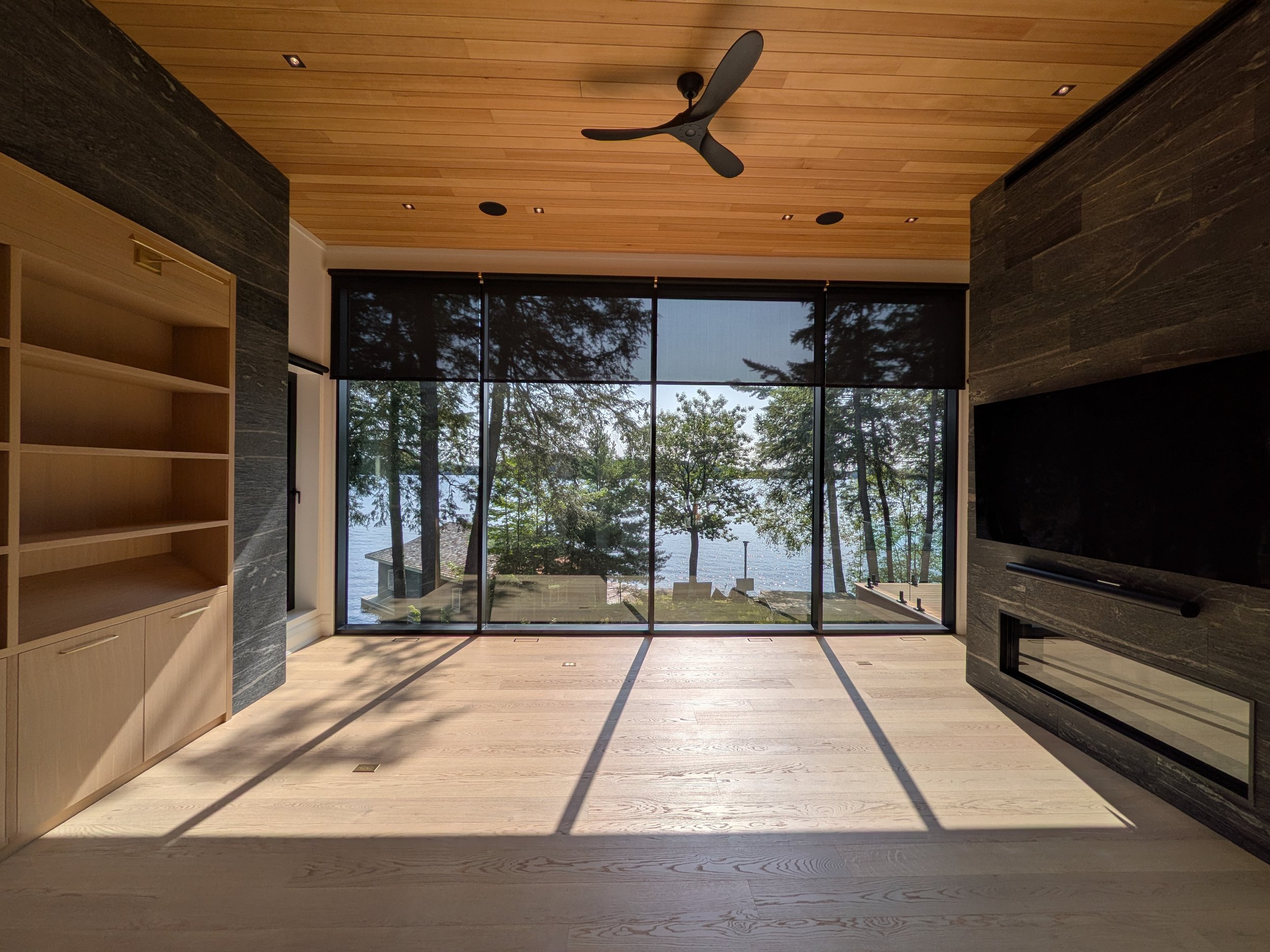 Empty living room with large glass windows showing a lakeside view, light wood flooring, wooden ceiling with ceiling fan, and dark stone walls with a mounted TV and fireplace.