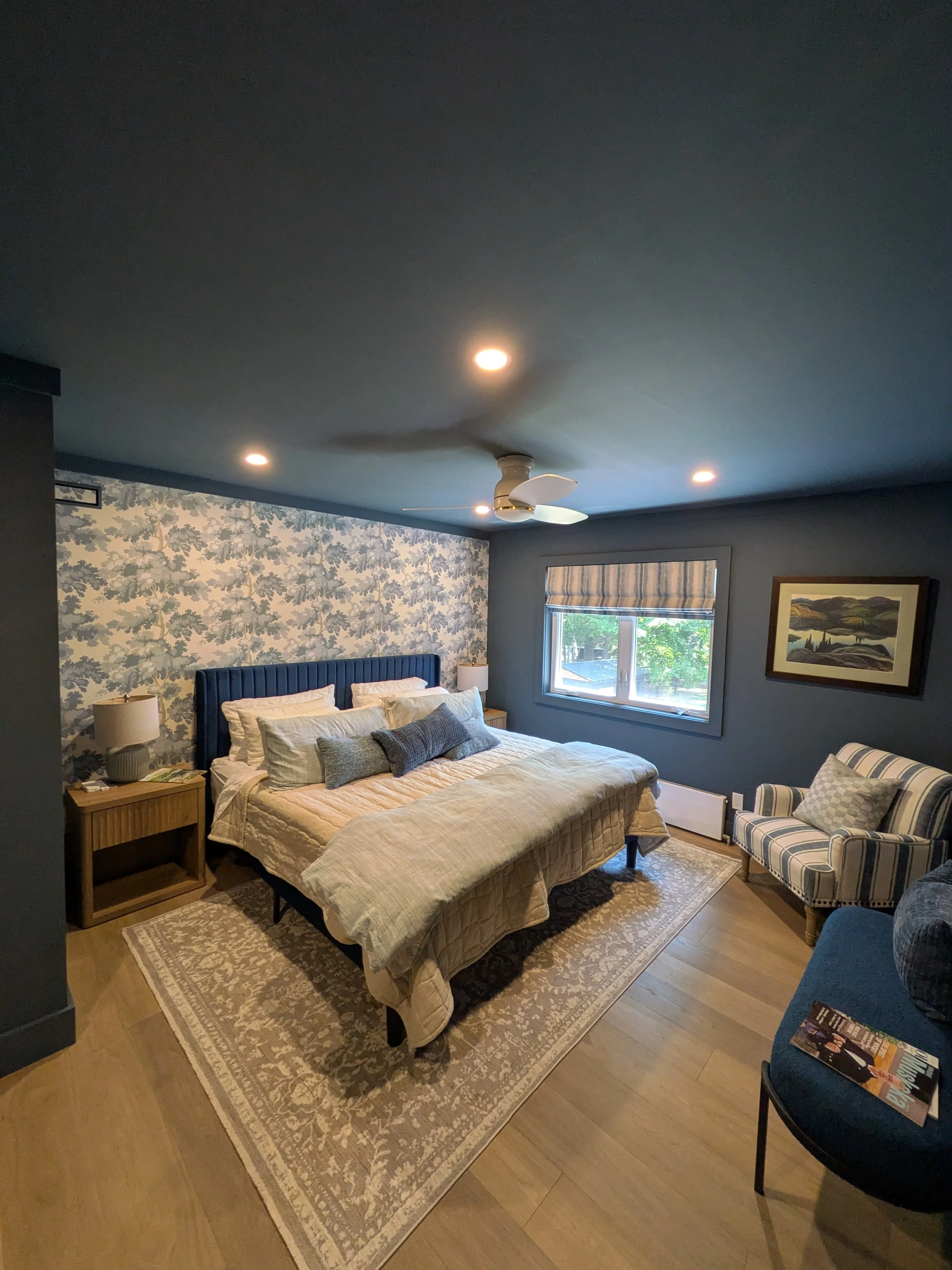 A cozy bedroom with a large bed, a striped armchair, and a window with a striped shade. The room has a patterned wallpaper accent wall, hardwood flooring, and modern lighting with recessed ceiling lights.