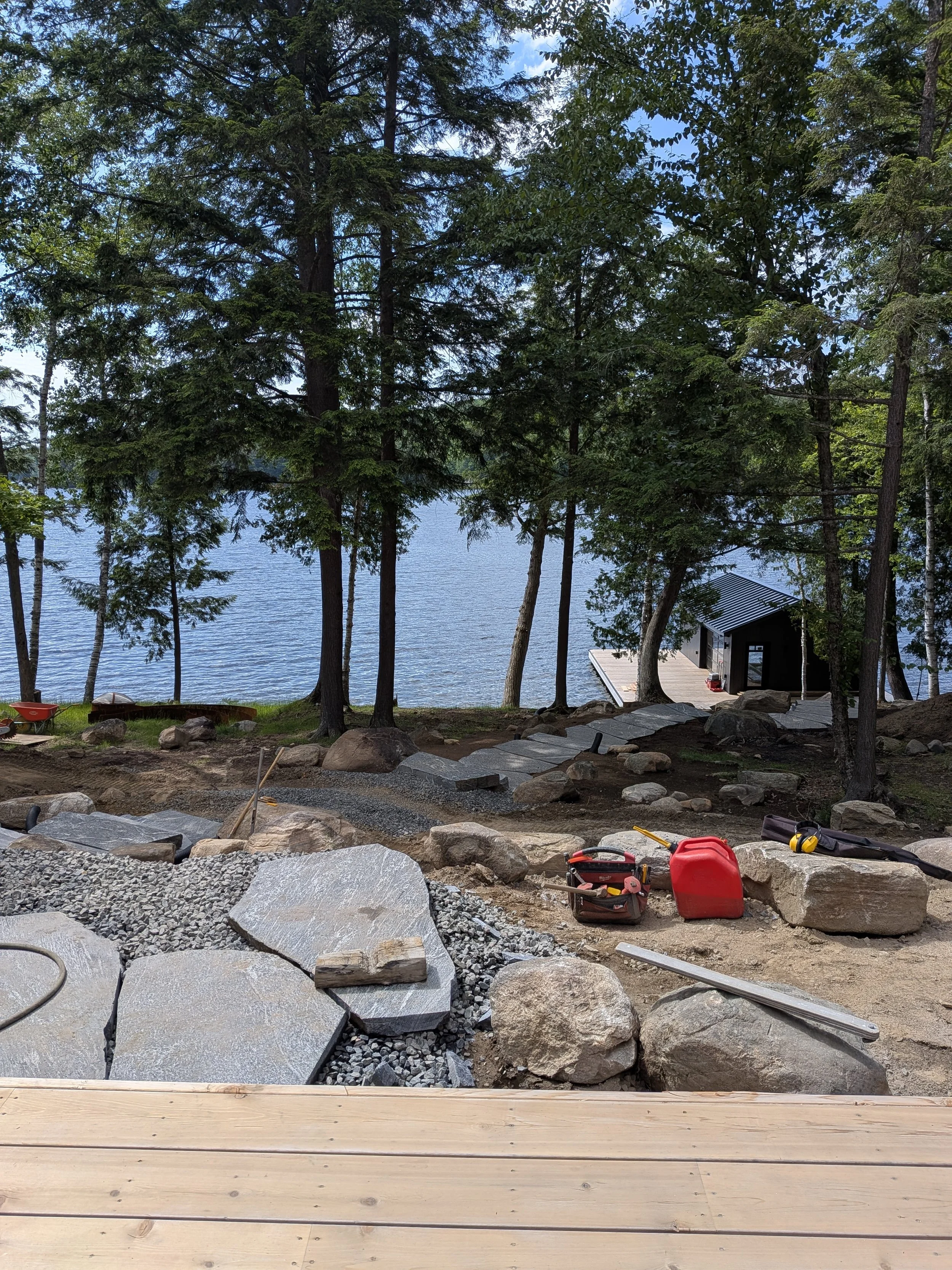 Construction site near a lake with large rocks, tools, and a partially built wooden deck. Trees and a small black building are visible in the background.