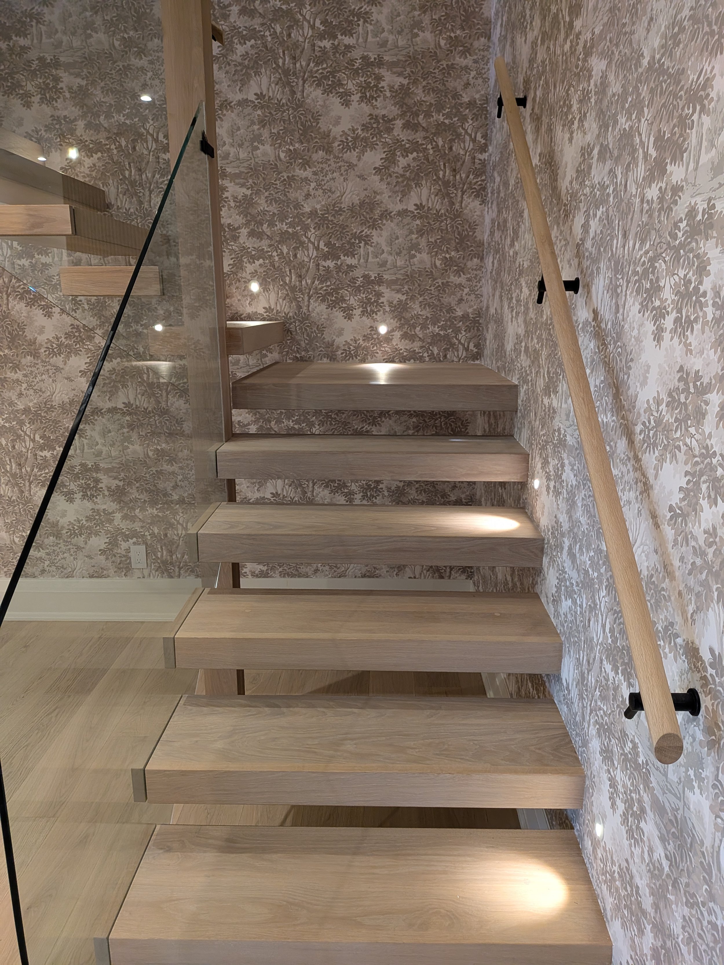 Wooden staircase with glass railing and a handrail on the right side, marble-patterned wallpaper, and small embedded lighting.