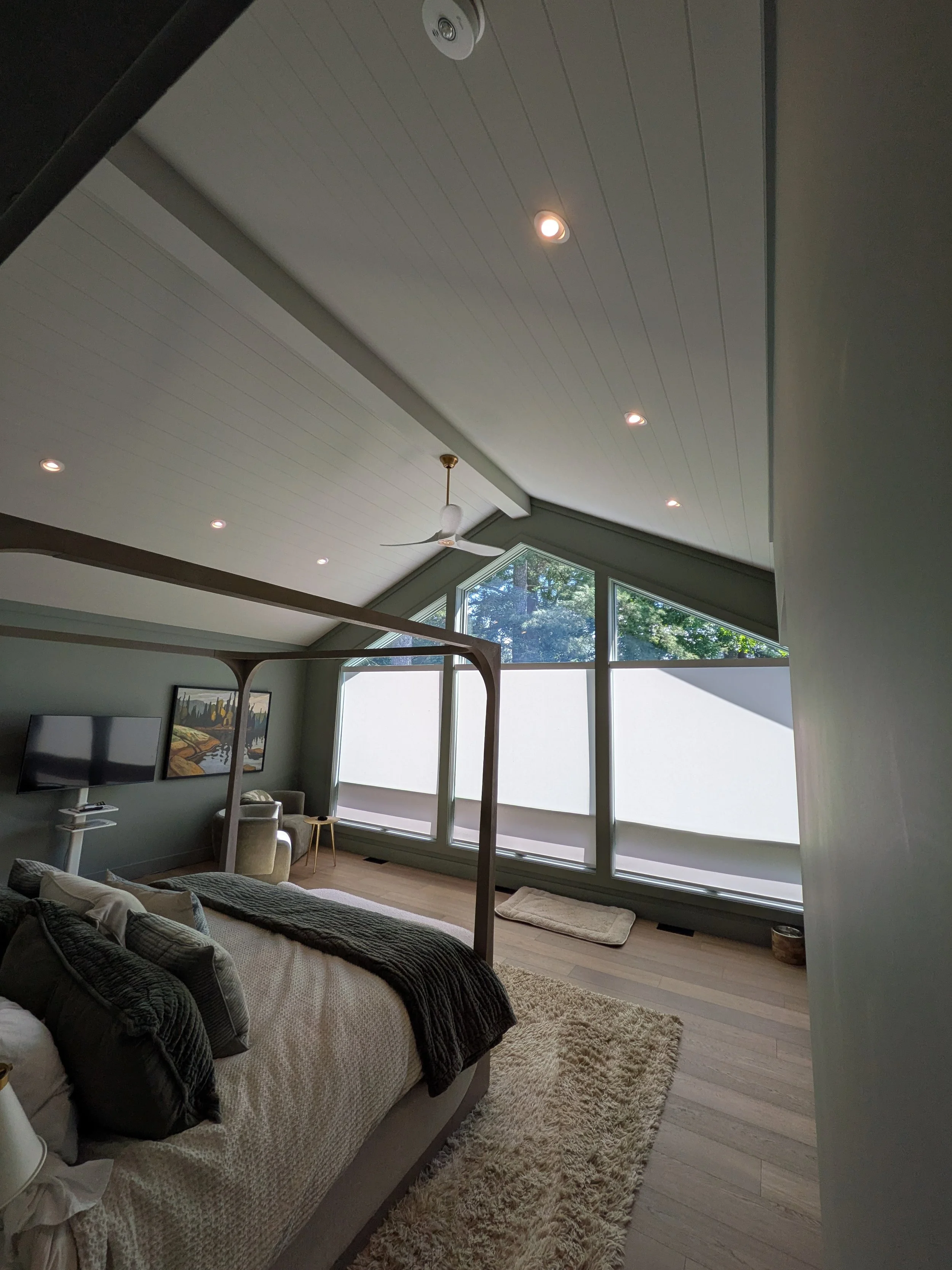 A bedroom with a sloped ceiling, a canopy bed, a flat-screen TV, a painting, a chair, a ceiling fan, and large windows with blinds.