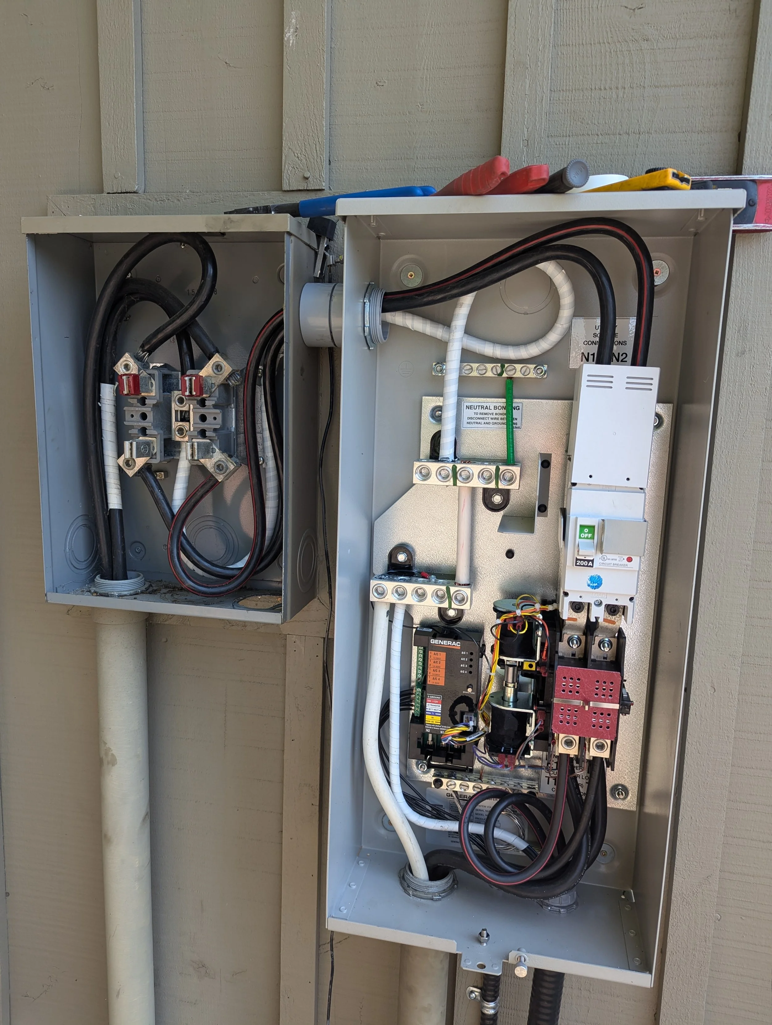 Open electrical box with wiring, circuit breakers, and a generator component inside, mounted on a wooden wall.