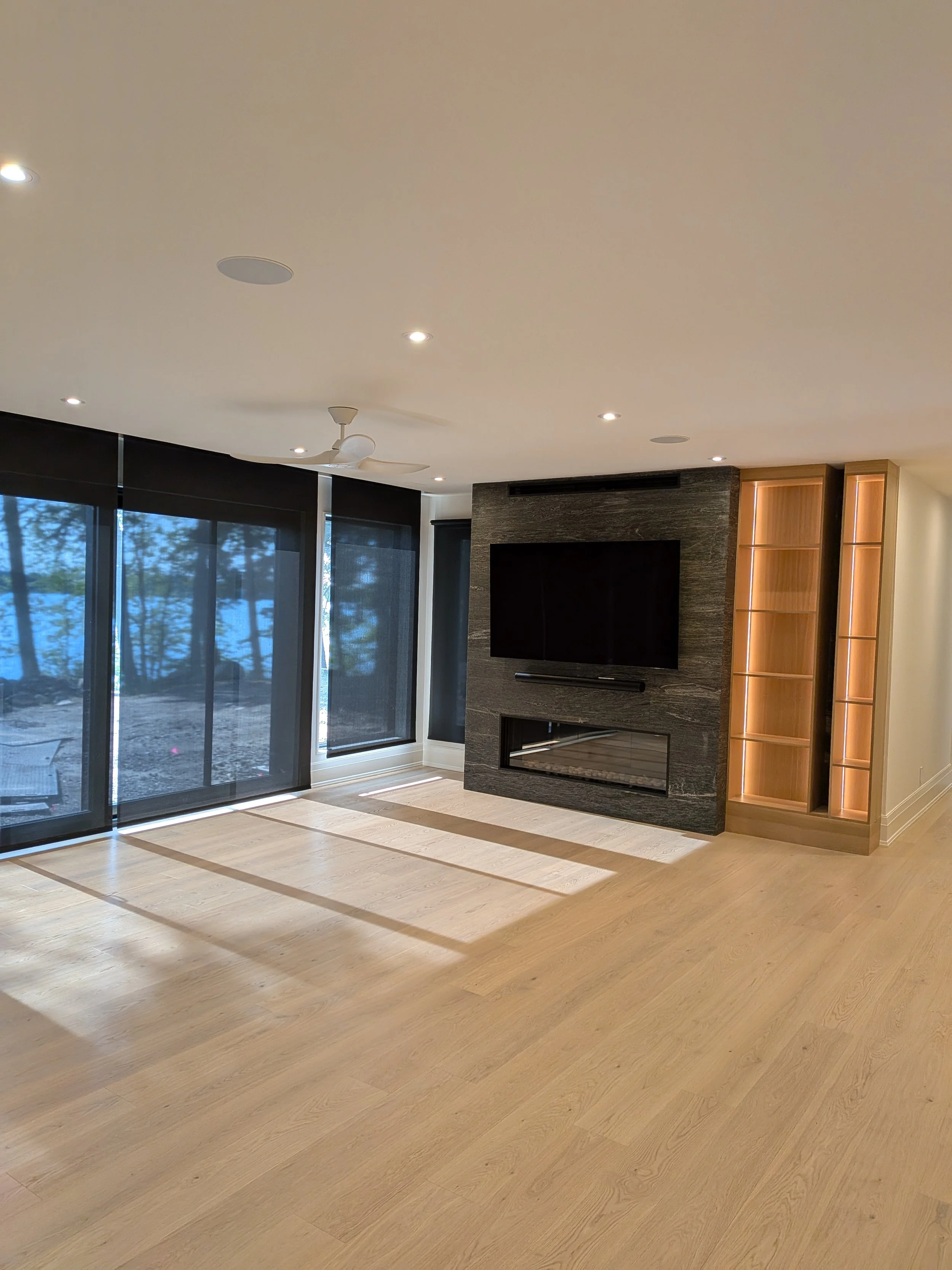 Empty living room with large windows, wooden flooring, and a black stone fireplace with a mounted TV above. Built-in wooden shelving with lighting on the right side.
