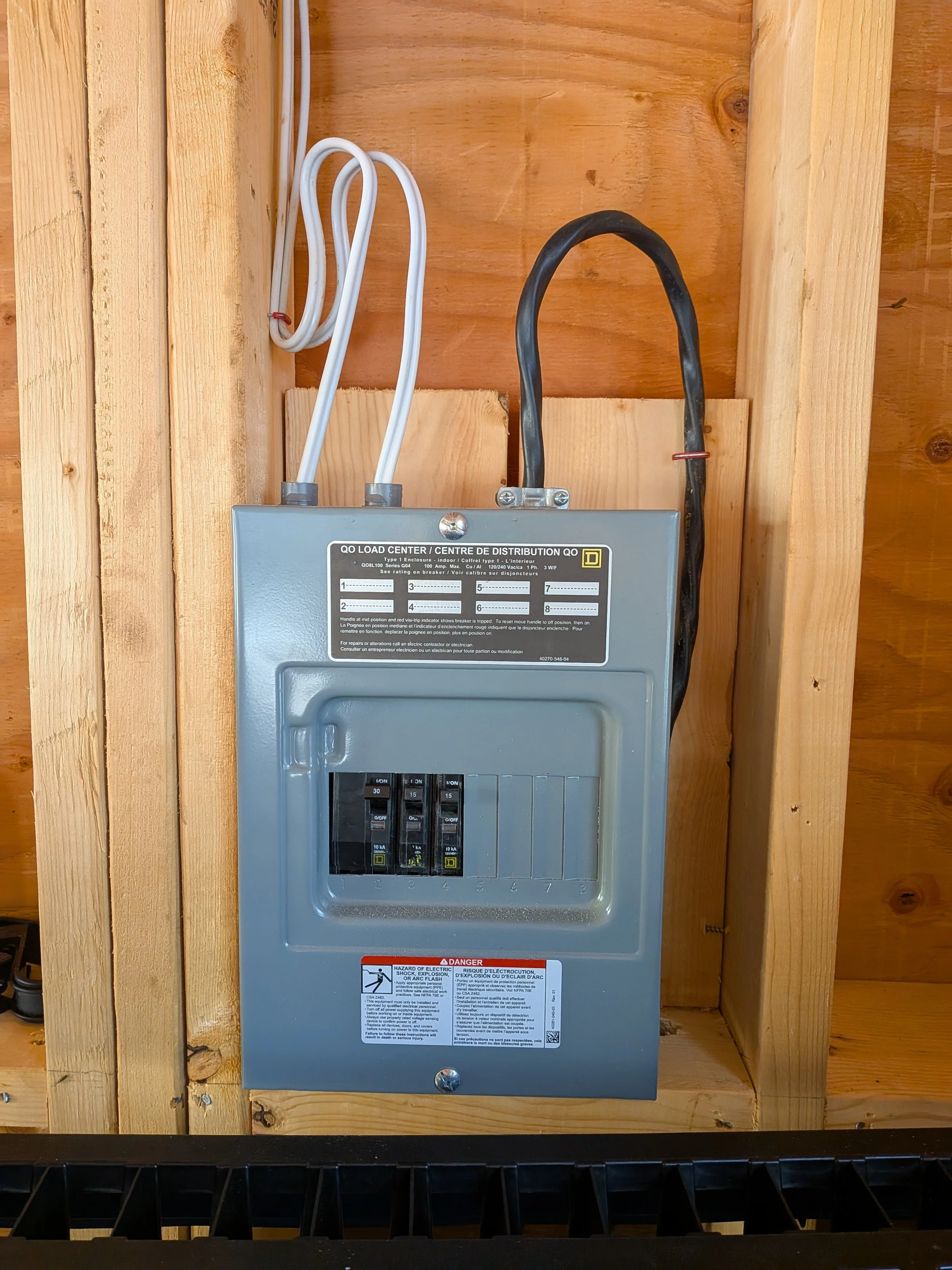 Electrical circuit breaker box mounted on a wooden wall with white and black wires connected to it.