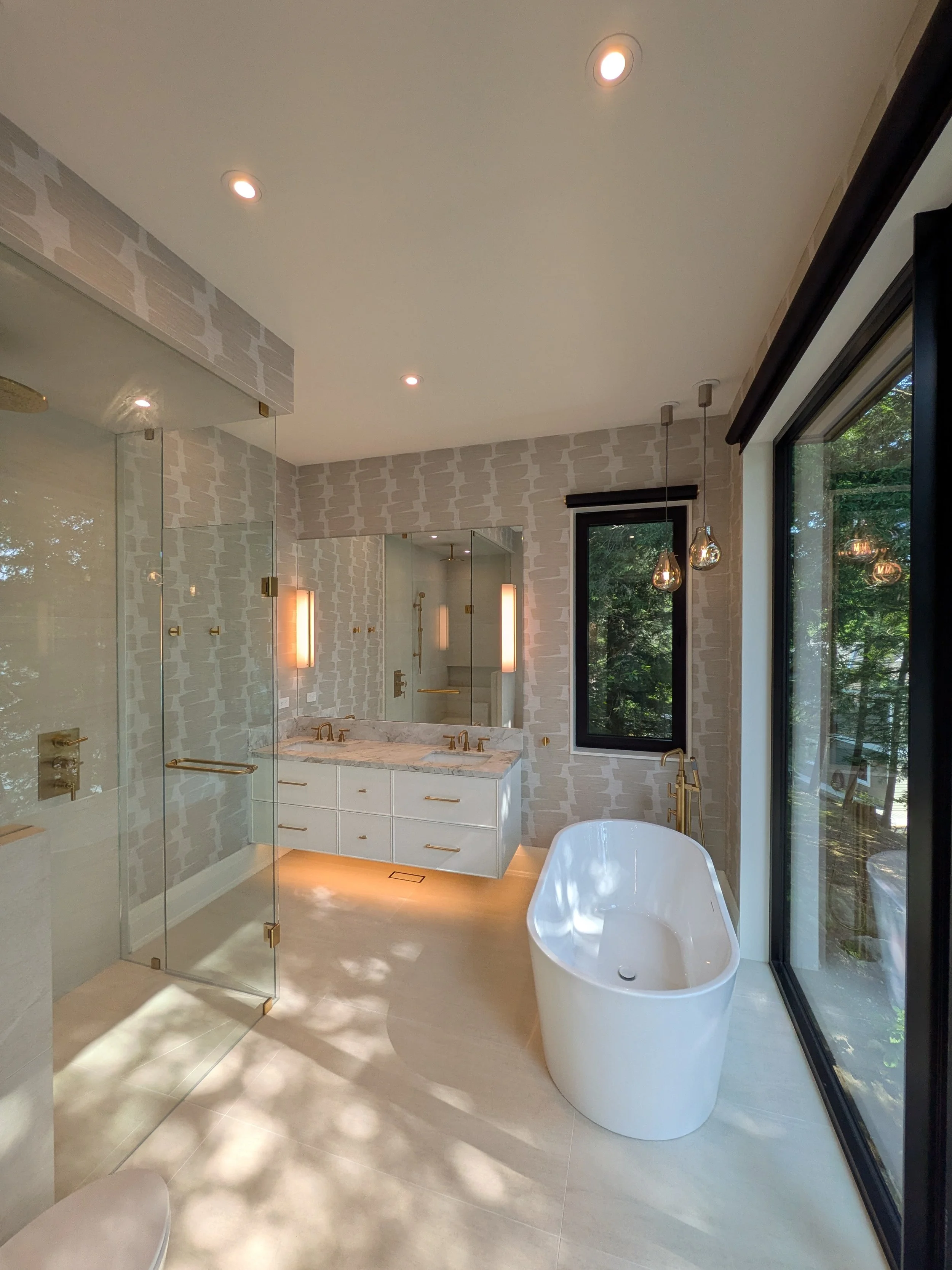 Modern bathroom with a freestanding bathtub near glass sliding doors, double vanity with gold fixtures, shower with glass enclosure, and large windows showing outdoor greenery.