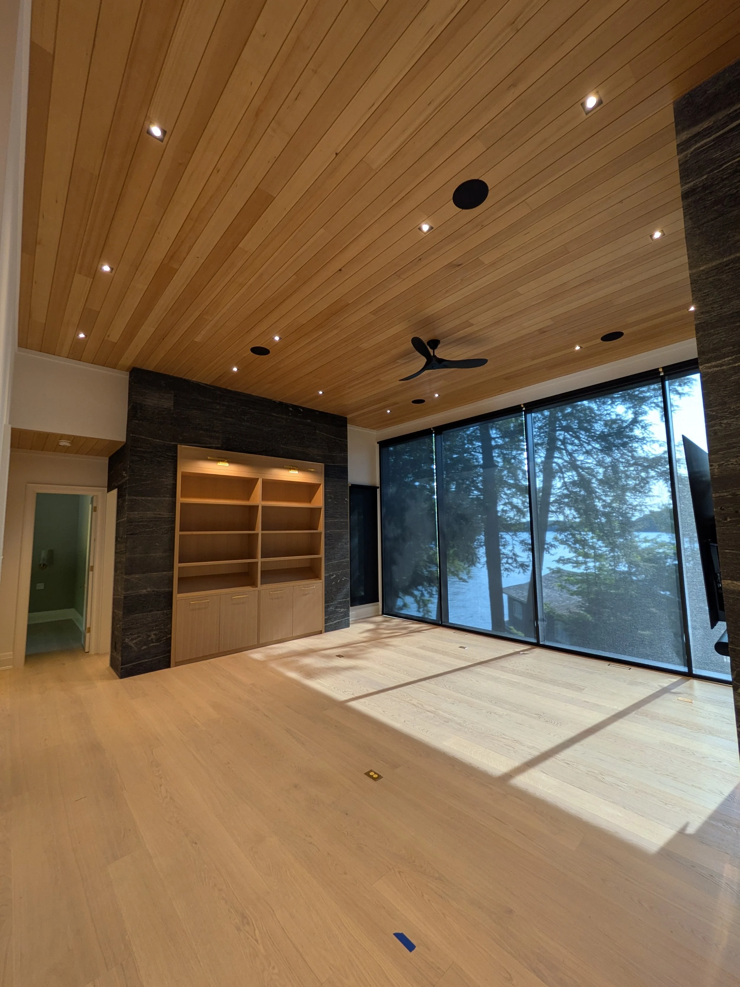 Empty living room with wooden flooring and ceiling, built-in bookshelf, large glass windows with tree view, ceiling fan, and recessed lighting.