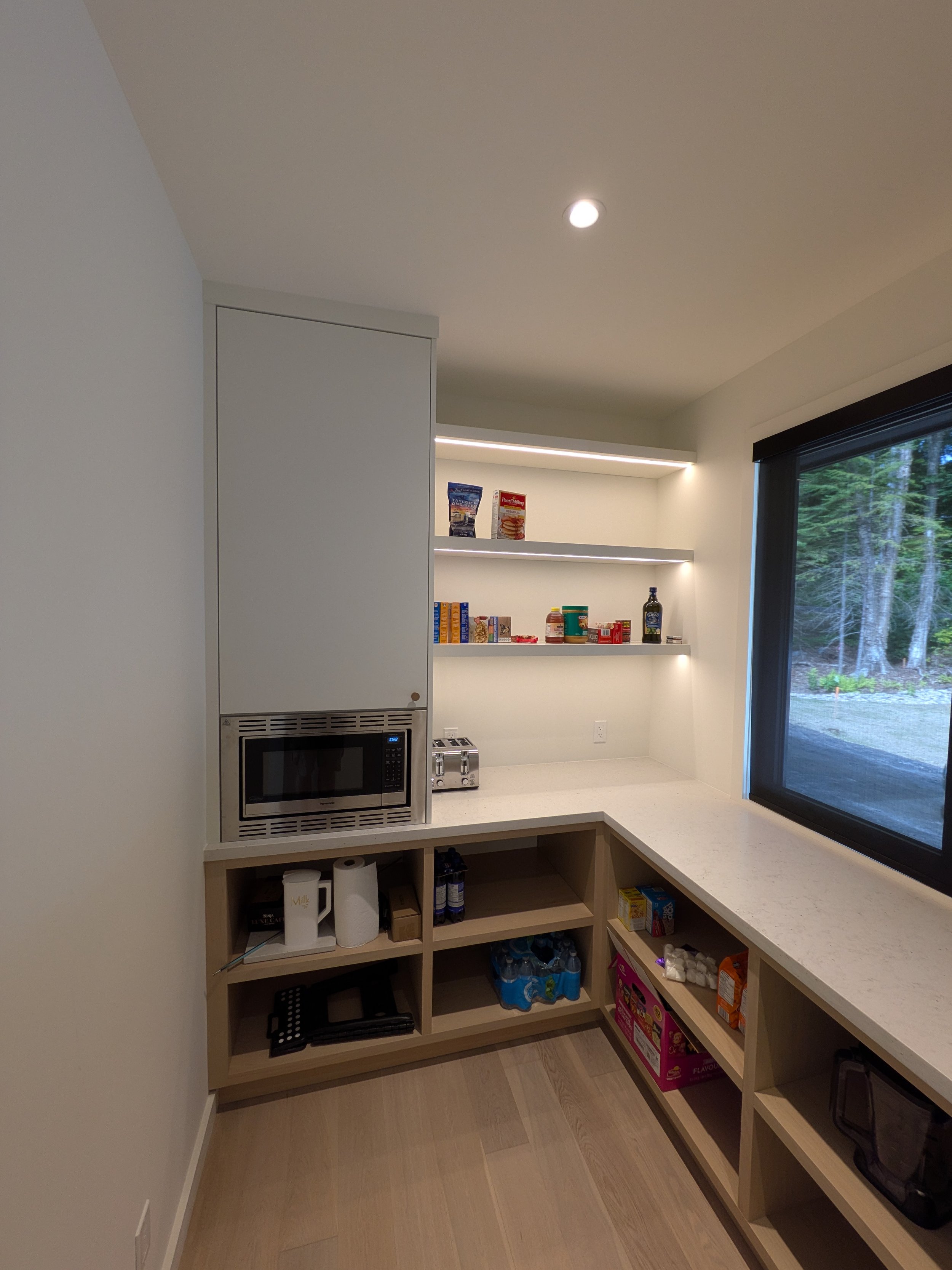 Modern kitchen pantry with open shelving, a mini microwave, and a large window showing trees outside.