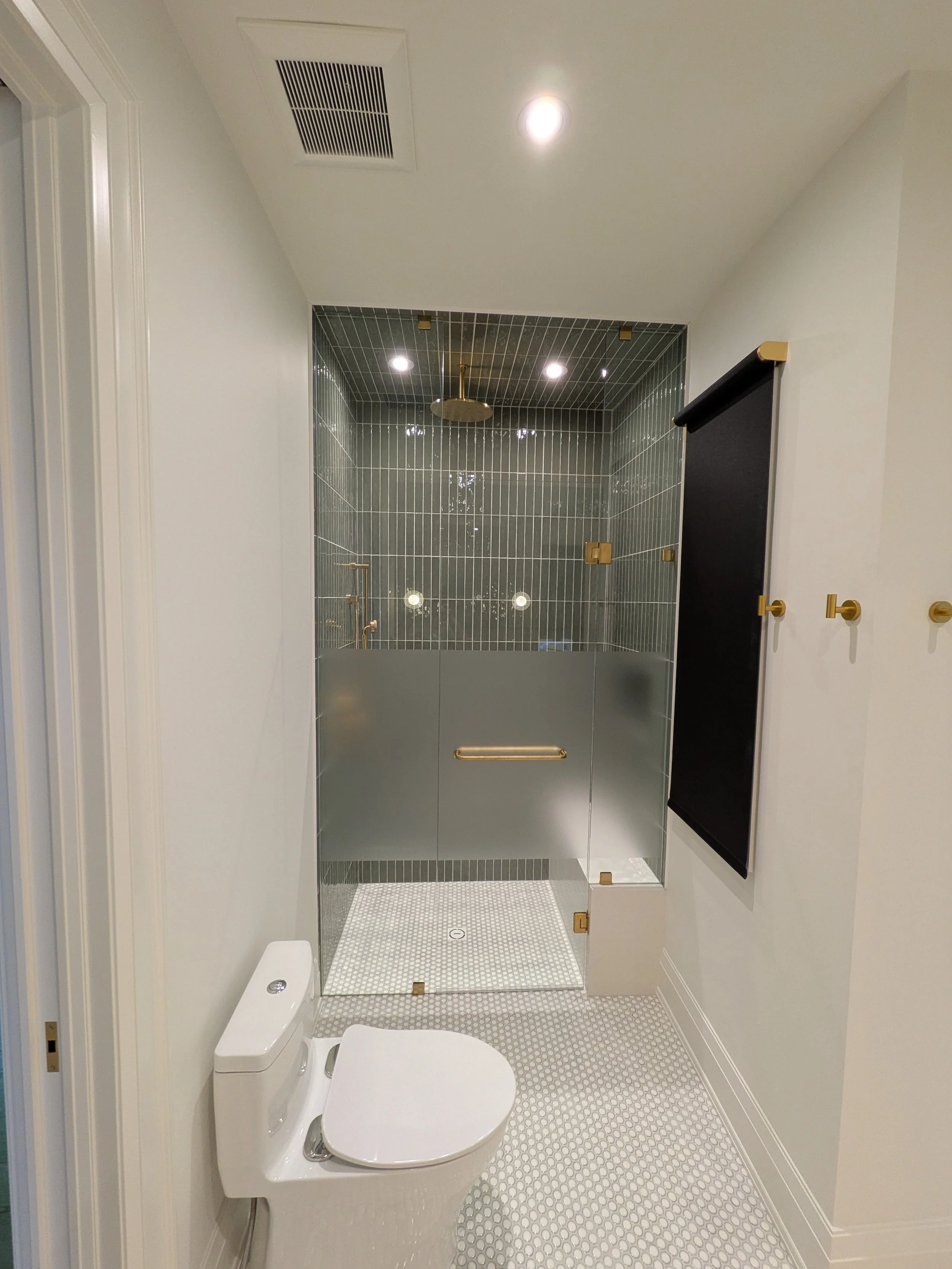 Modern bathroom with a walk-in shower with glass enclosure, gold fixtures, a white toilet, and a black roller shade on the wall.