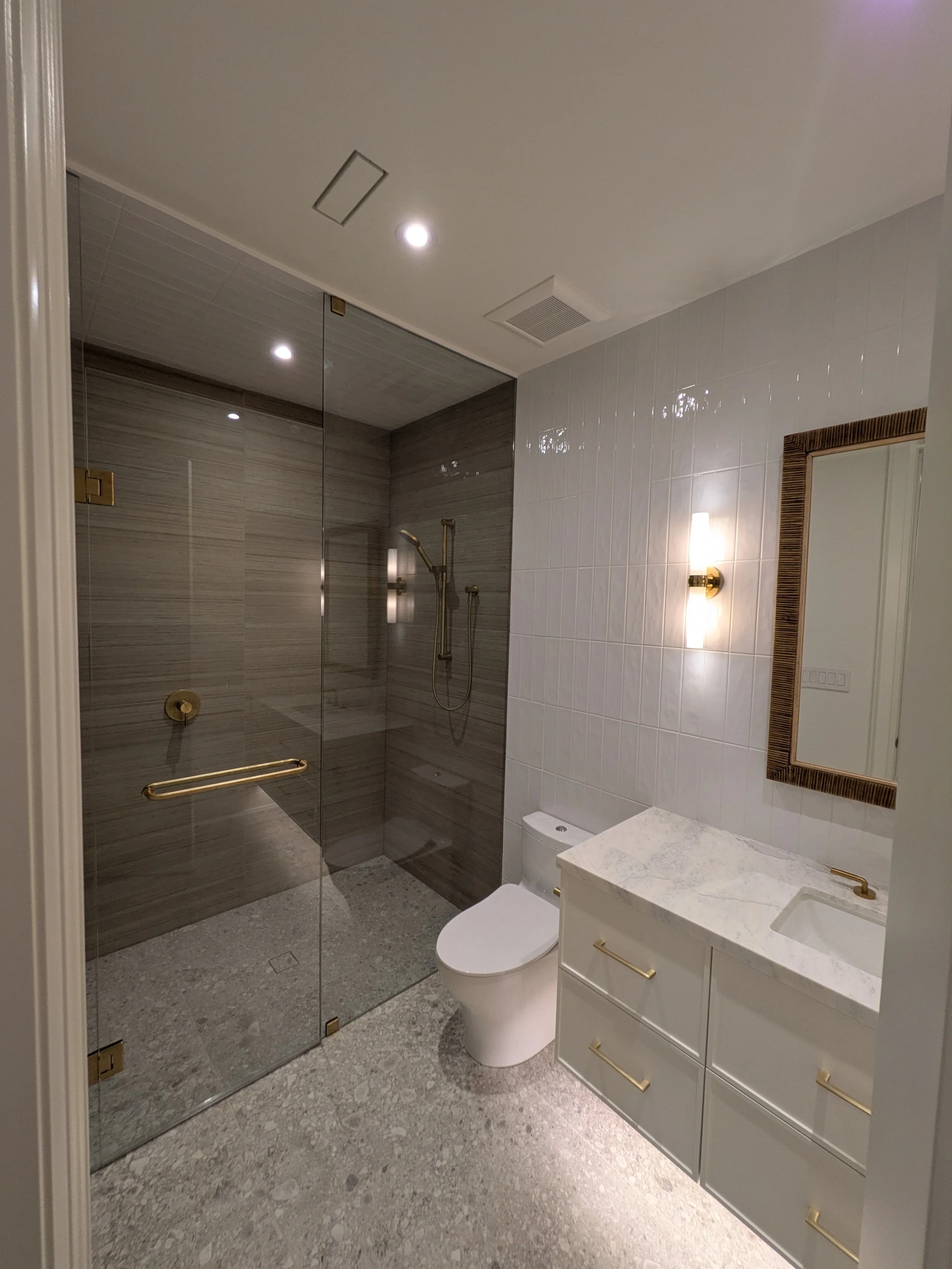 Modern bathroom with glass shower enclosure, white toilet, and white vanity with marble countertop and brass handles. Wall mirror and brass light sconce. Gray tiled floor and white tiled walls.