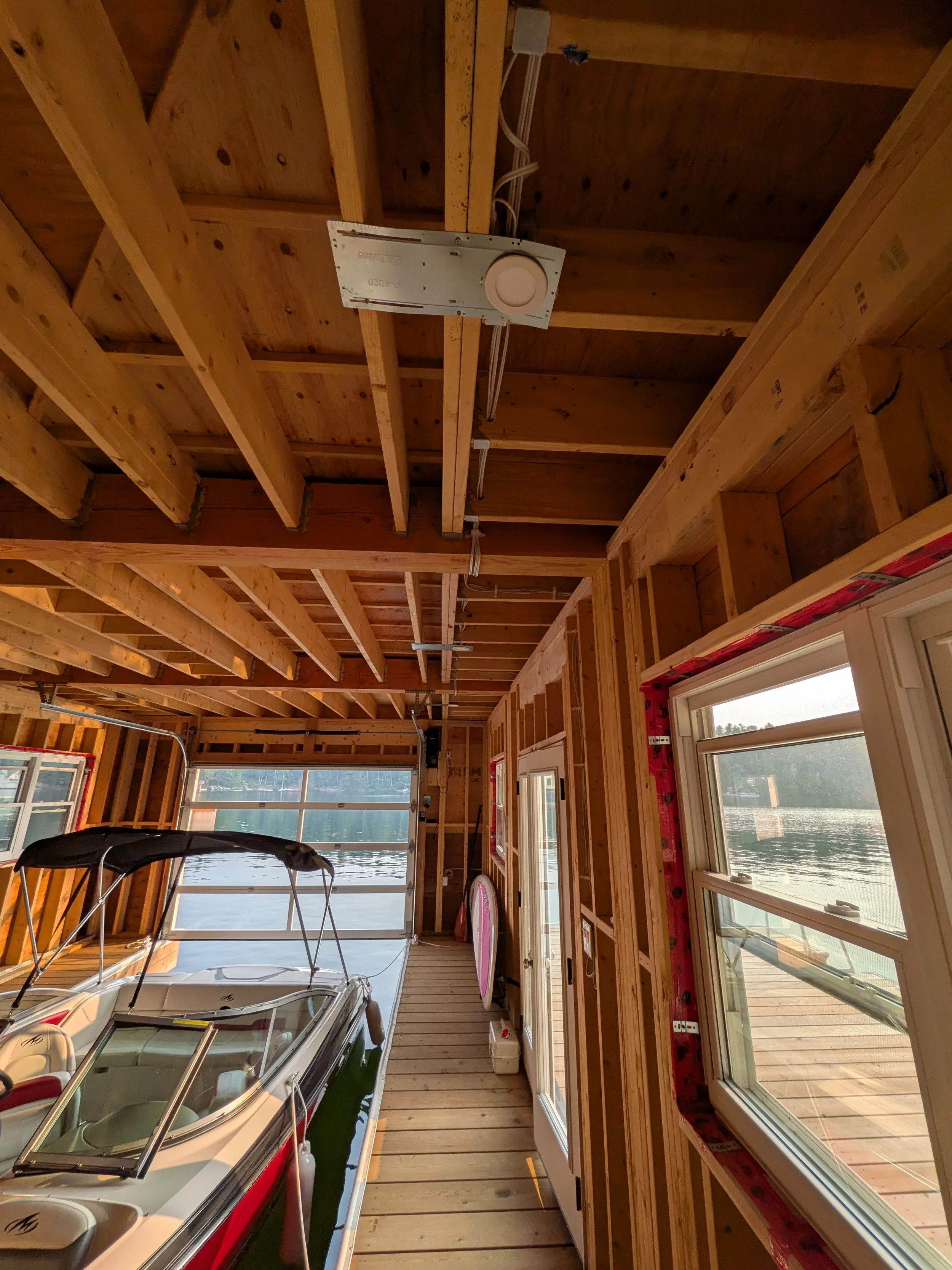 Interior of an unfinished boathouse, showing exposed wooden framing, windows looking out to a lake, and a boat parked inside.