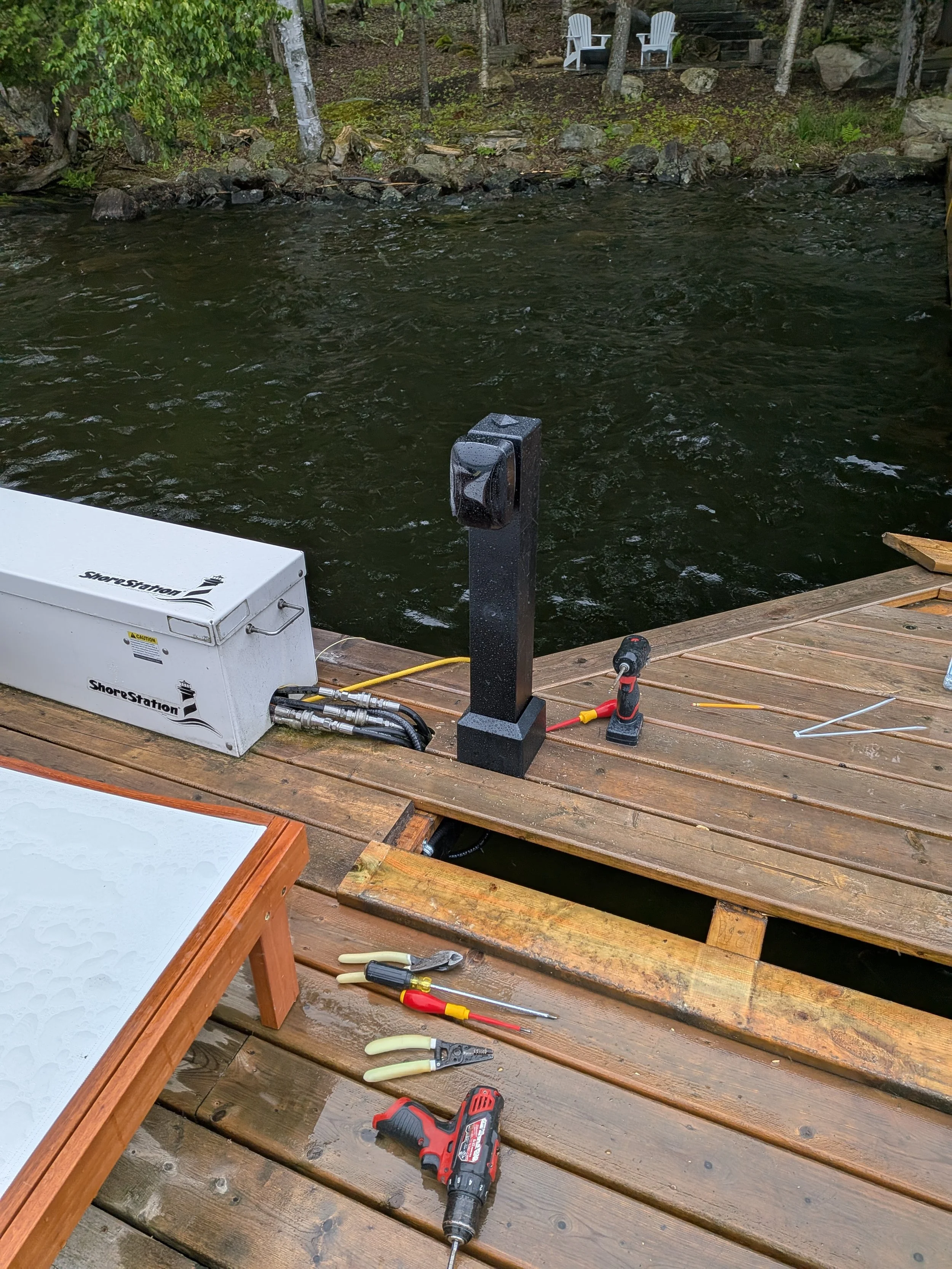 Tools and equipment on a wooden dock near water, with a black pole, a white ShoreStation box, and chairs in the background.