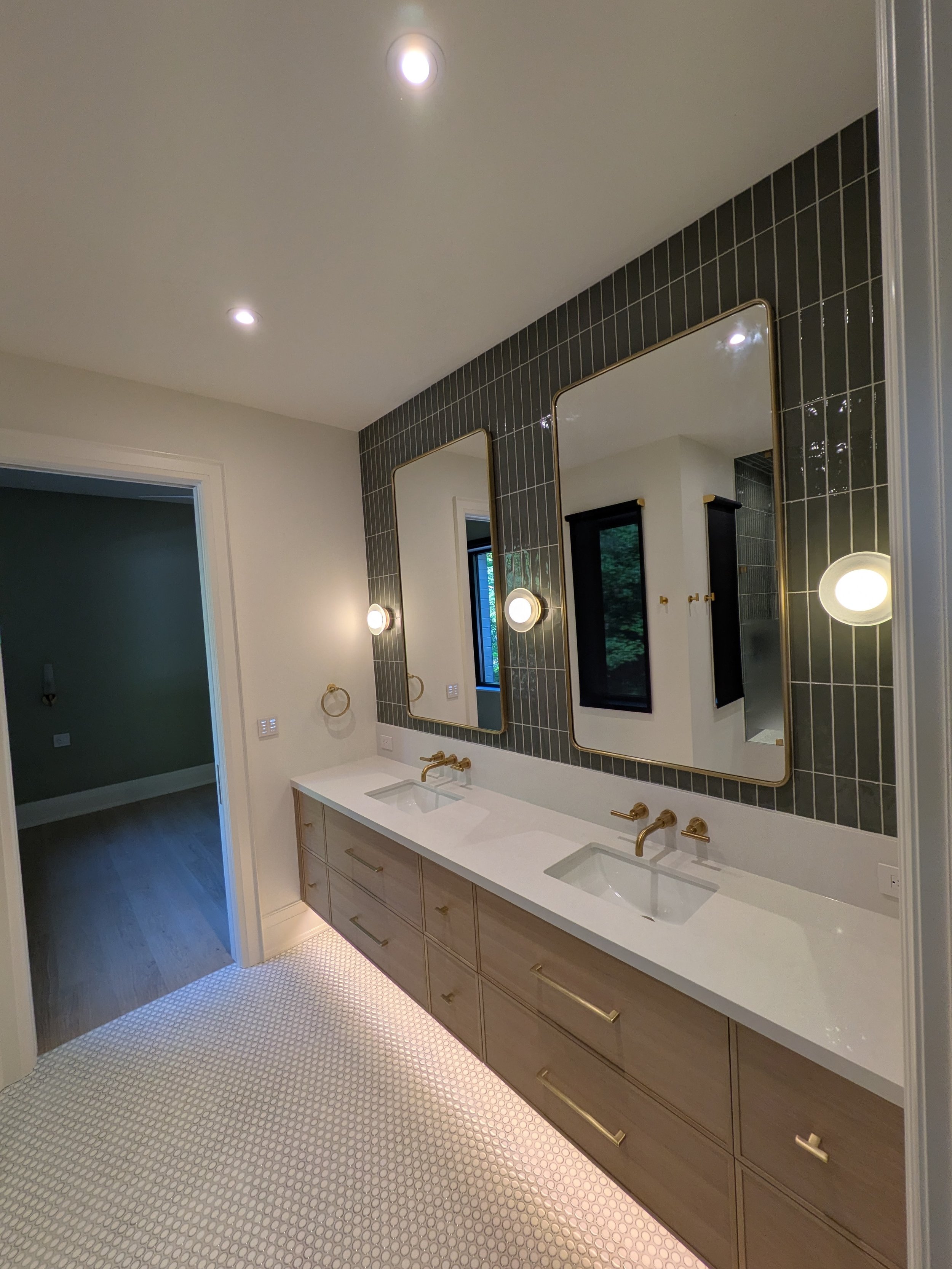 Modern bathroom with dual sinks, large mirrors, gold fixtures, black tiled wall, and white mosaic floor.