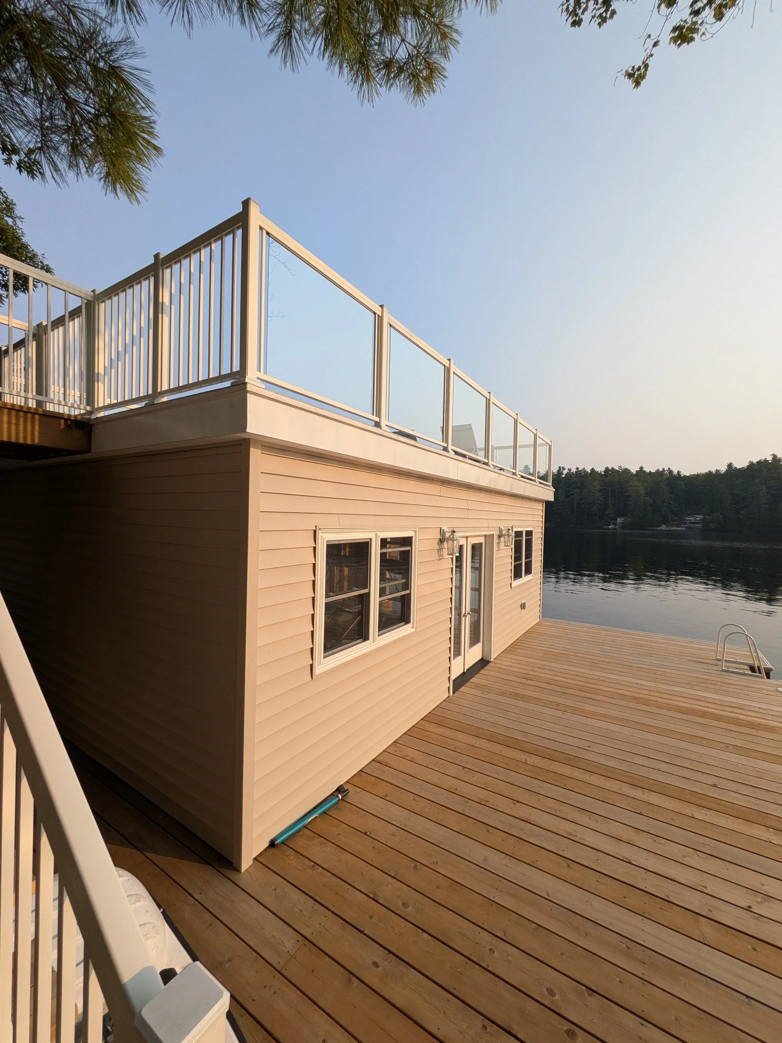 A two-story boathouse built on a wooden dock extending over a Lake Joseph, with a balcony on the upper level and trees in the background.