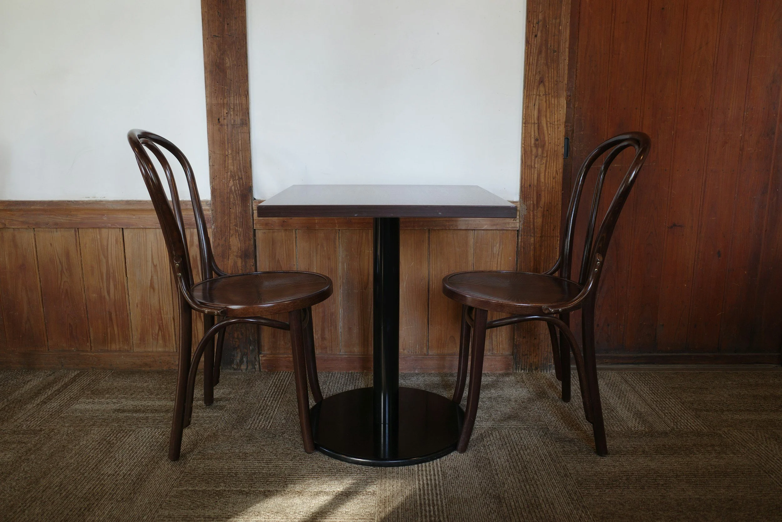 Two wooden chairs facing each other with a small square table in between, in a rustic wood-paneled room.