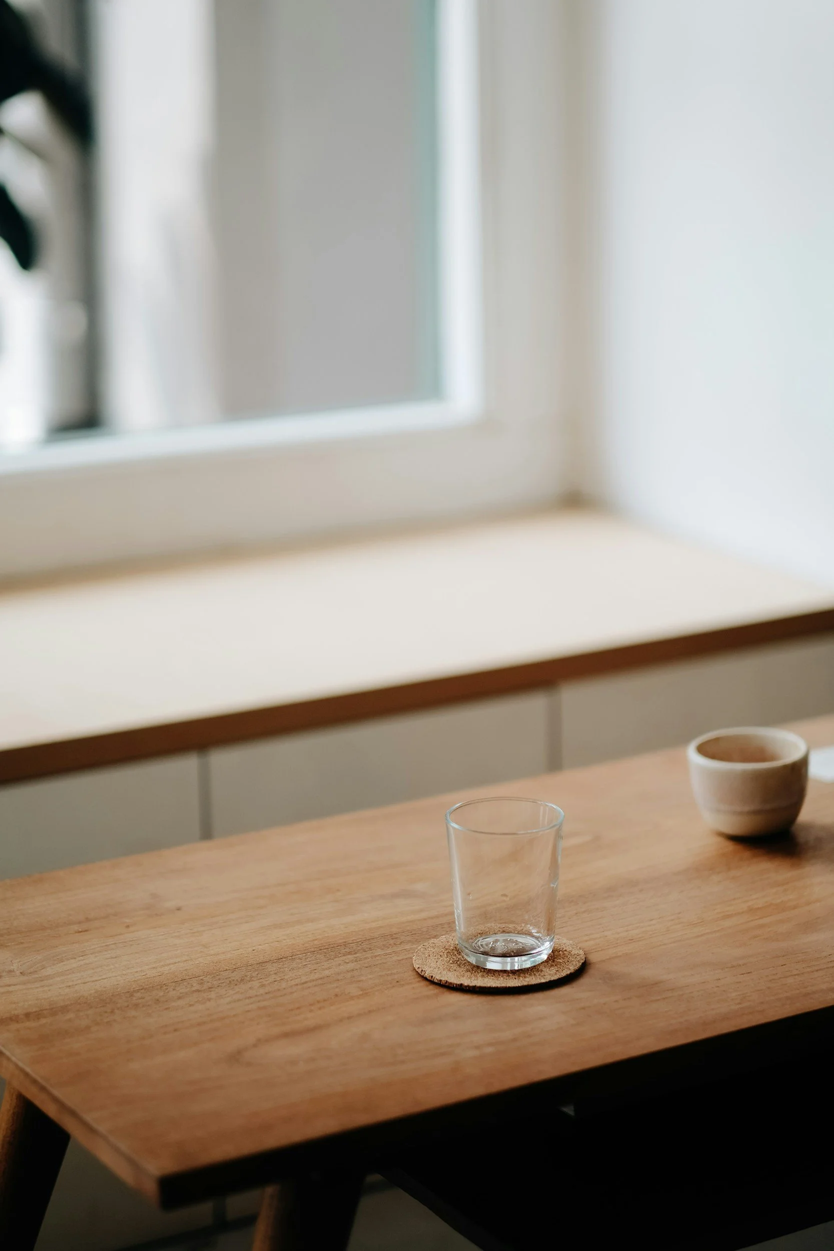 A clear glass on a cork coaster and a small beige bowl on a wooden table near a window with white light.