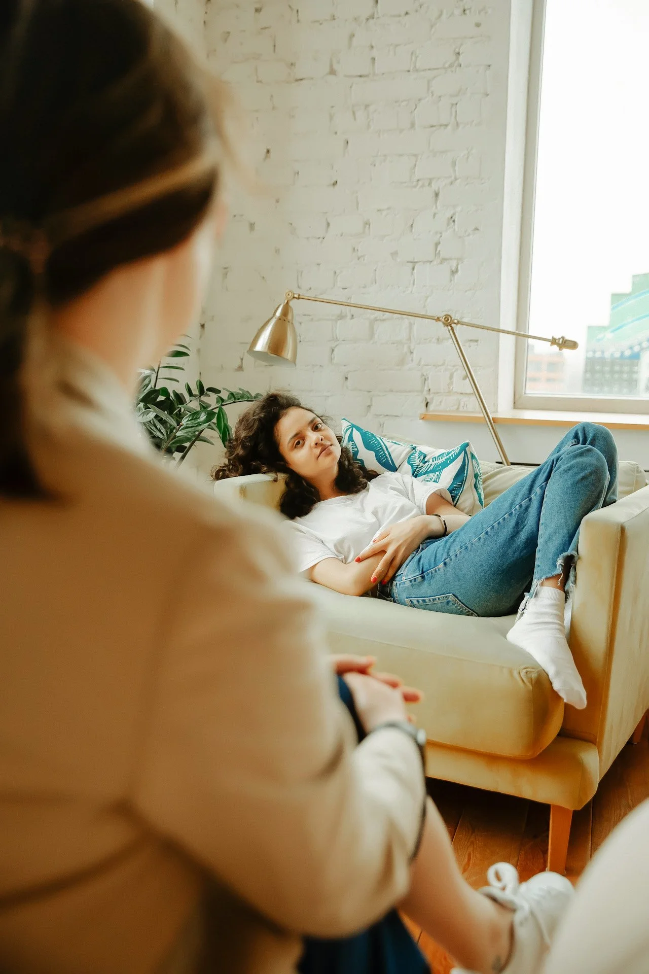 A young woman lying on a sofa and looking at another woman sitting nearby in a bright living room with a white brick wall and a large window.