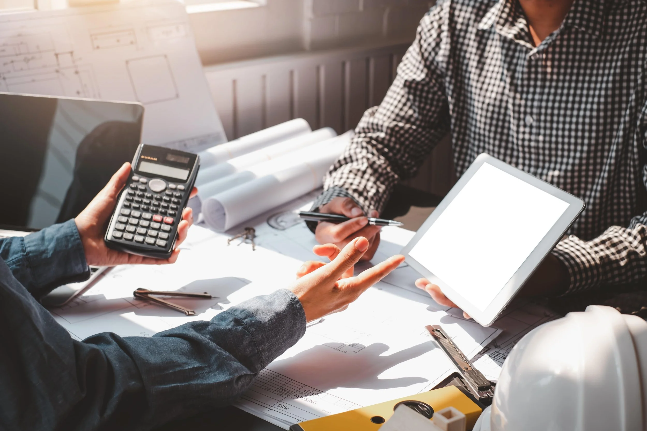 Two people reviewing architectural plans with a calculator and digital tablet, on a desk with blueprints and a white hard hat.