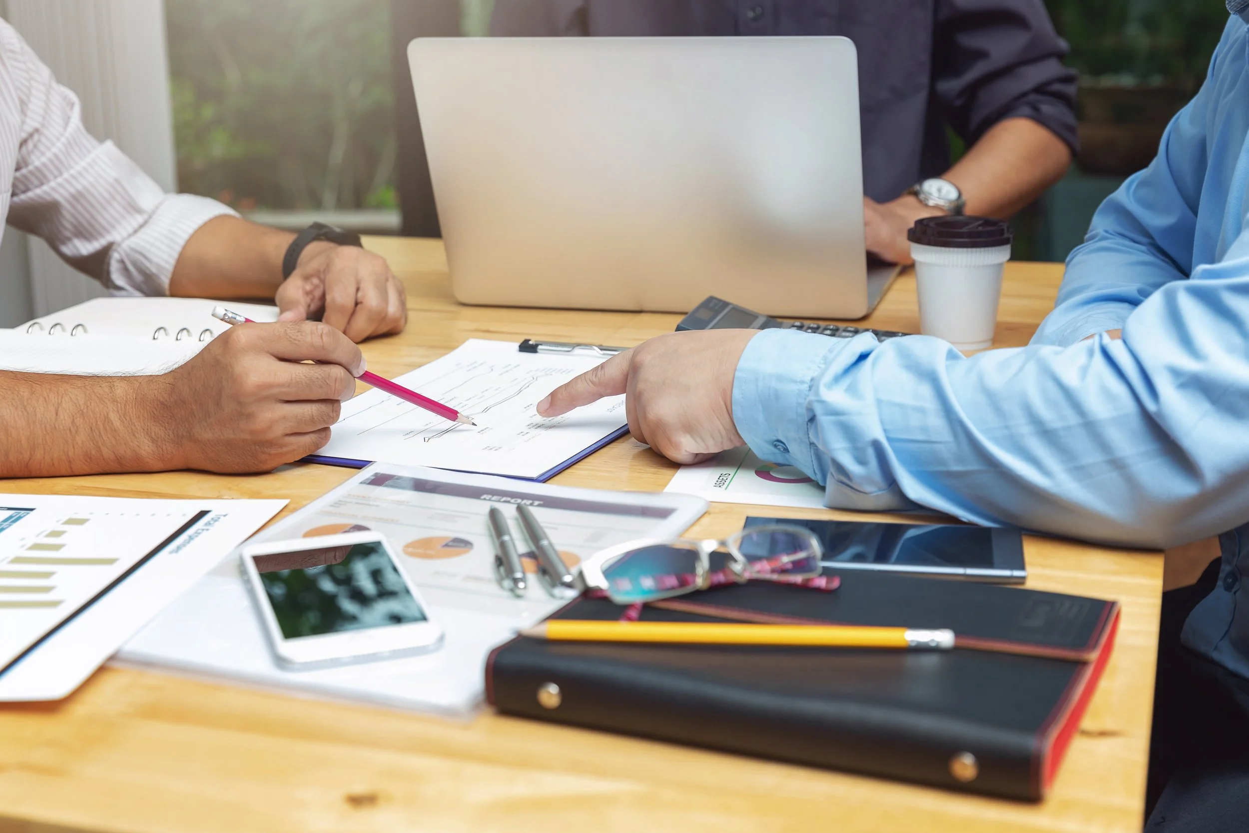 Three people working together at a wooden desk with laptops, documents, pens, a smartphone, and a coffee cup.