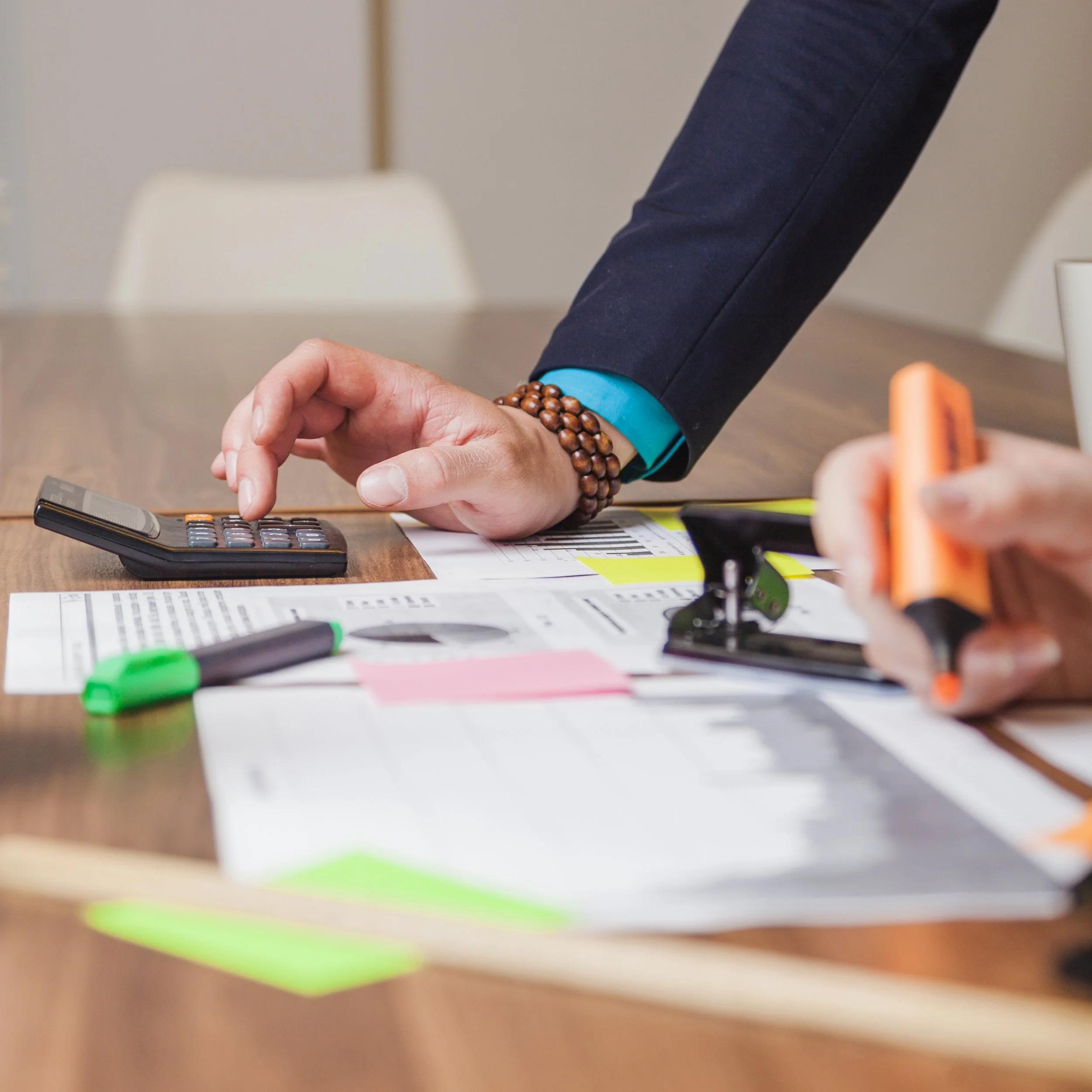 Person in business attire using a calculator at a cluttered desk with papers, highlighters, and office supplies.