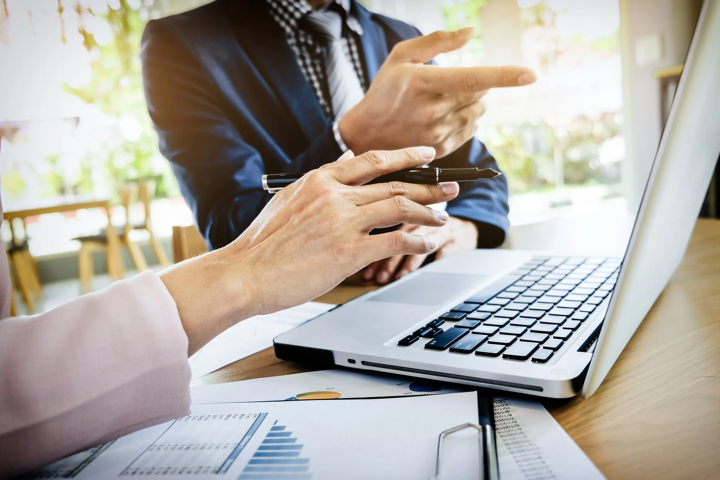 Two people in business attire having a discussion at a table with a laptop, papers, and a pen, in a bright, outdoor setting.