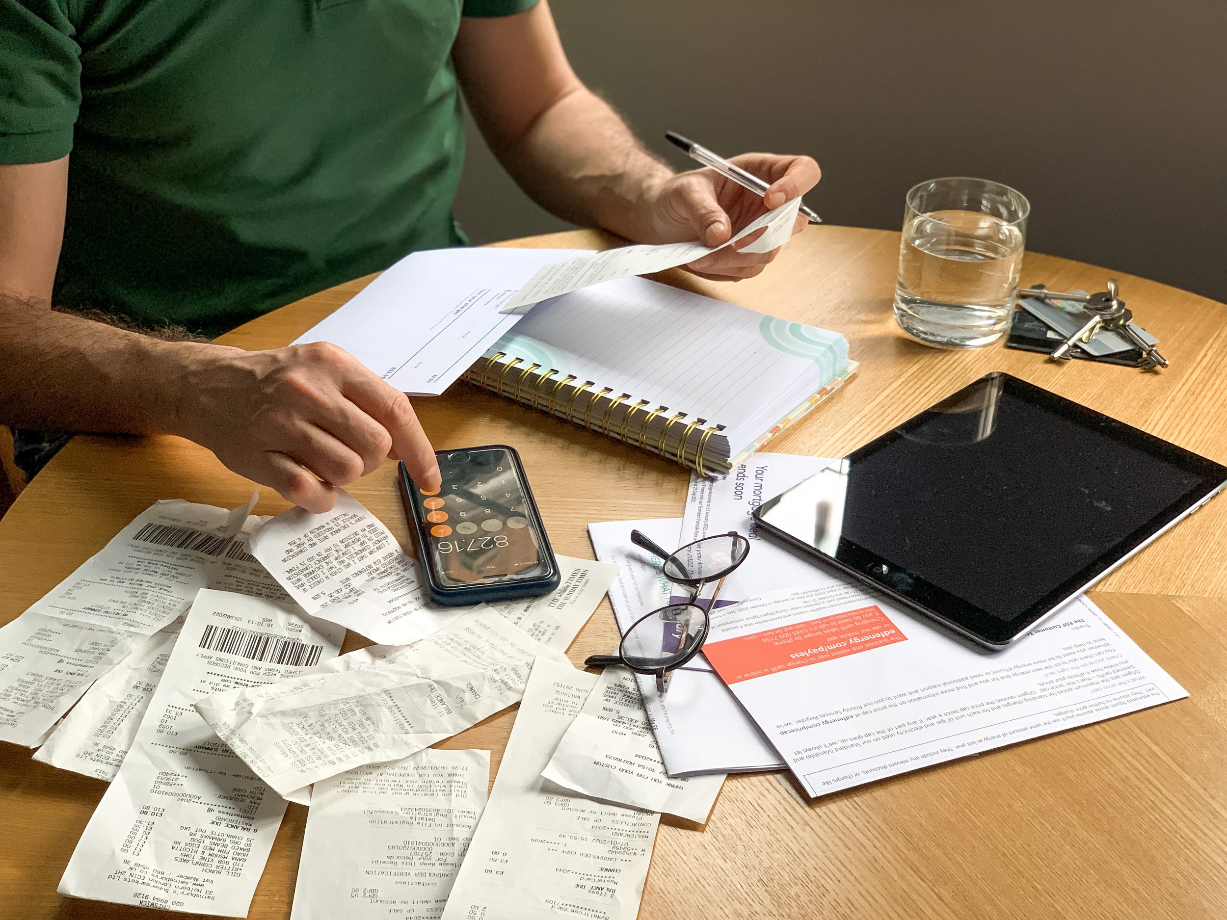 A person with a green shirt sitting at a wooden table cluttered with receipts, a calculator, a smartphone, glasses, a notebook, a tablet, and some documents, with a glass of water nearby.