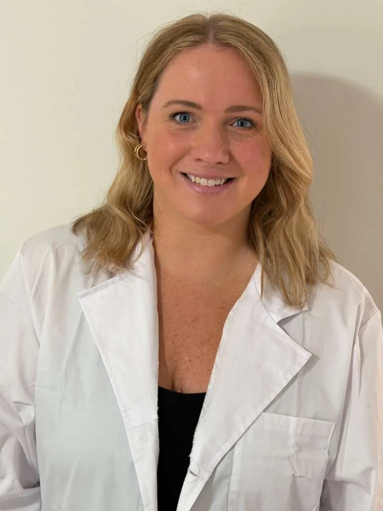 A woman with shoulder-length blonde hair, wearing a white lab coat and gold hoop earrings, smiling at the camera, standing against a plain light-colored background.