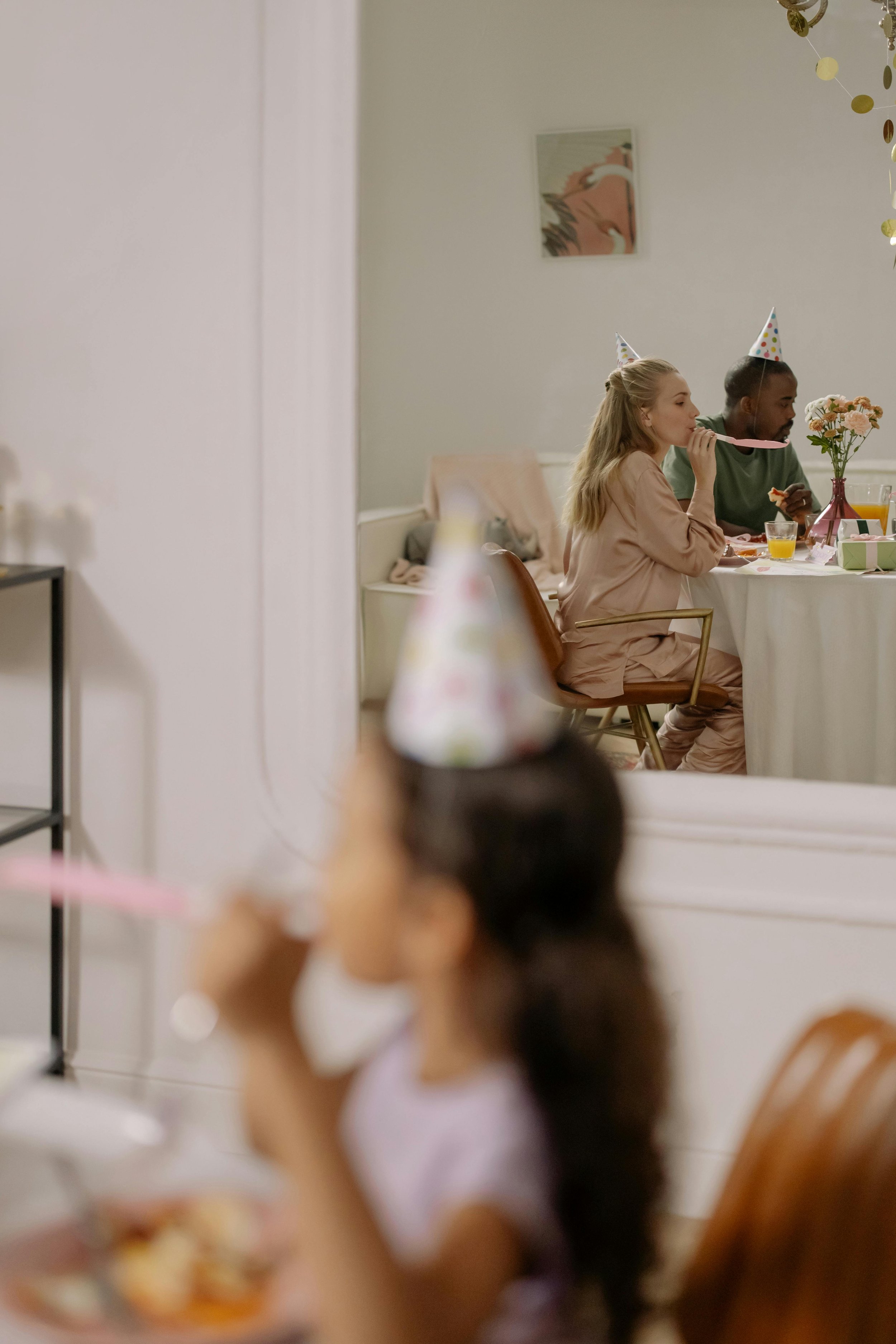 Bir çocuk masaya oturmuş doğum günü partisini kutluyor, önünde pasta ve parti şapkası var, diğer çocuklar ve yetişkinler masanın yanında kutlama yapıyor,  nice yaşlara