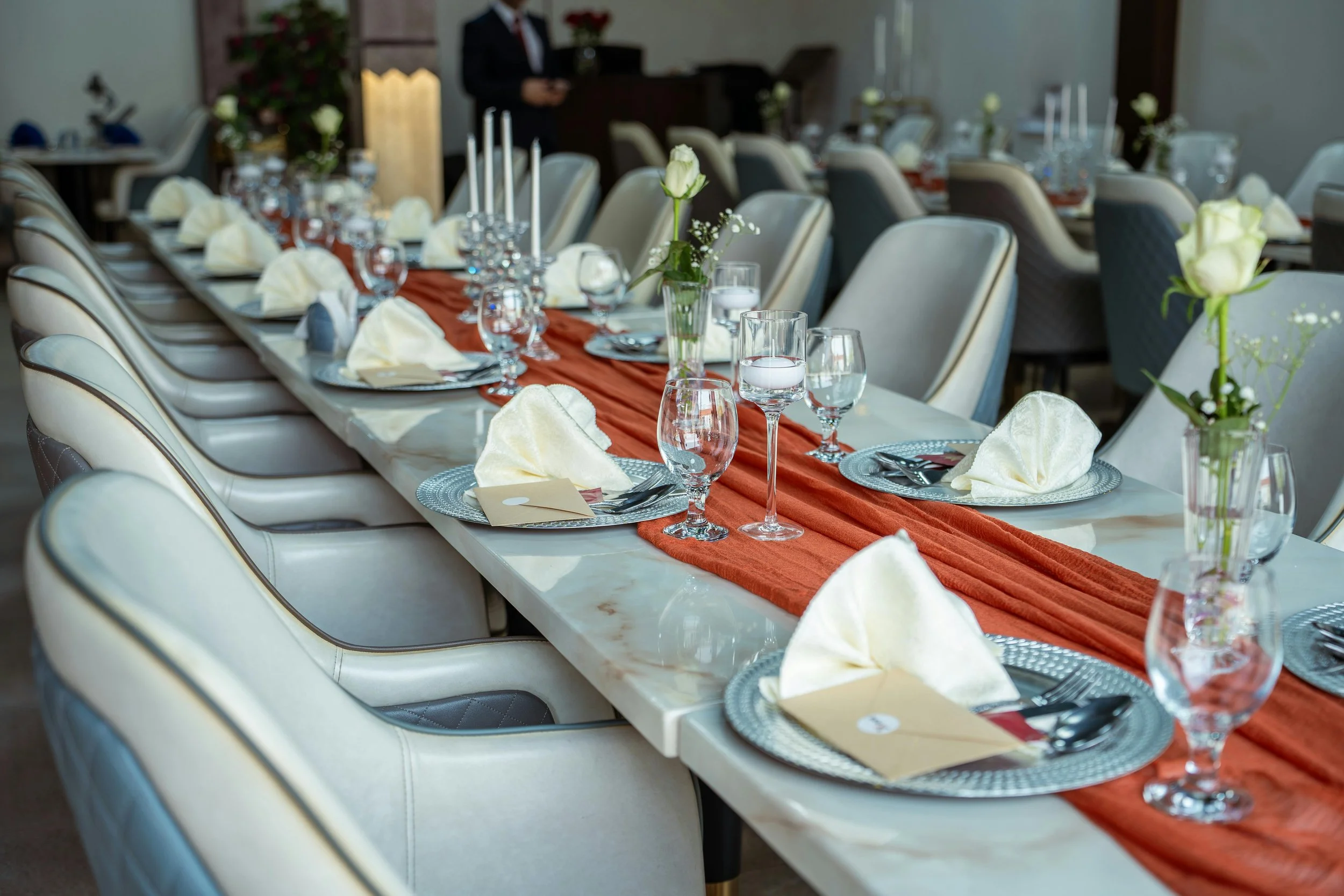 Şık bir masa düzeni, beyaz tabaklar, gümüş çatal ve bıçaklar, şeffaf su ve şampanya kadehleri, beyaz peçeteler, çiçeklerle süslenmiş, turuncu masa örtüsü üzerinde hazırlanmış.