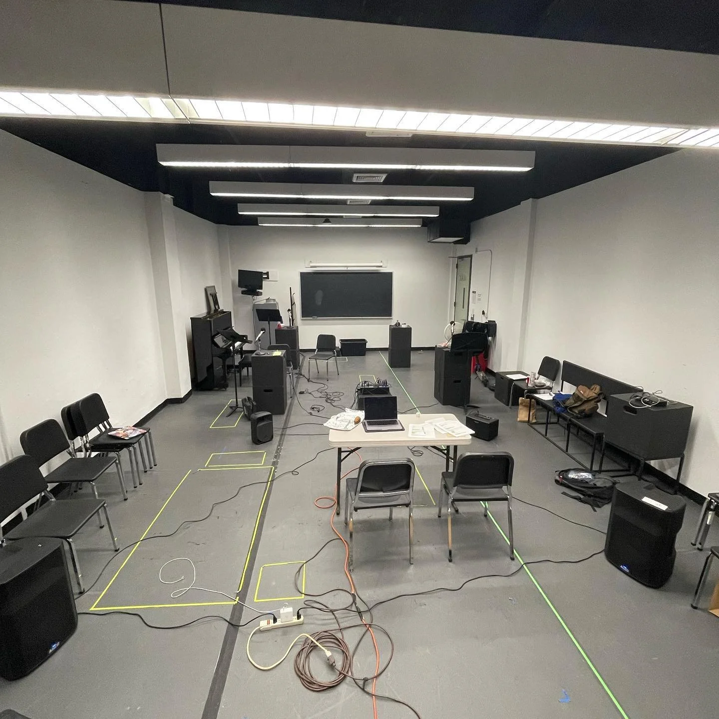 Empty classroom or conference room with chairs arranged in a semi-circle, electronic equipment including speakers, cables, a table with audio gear, and a blackboard at the front.