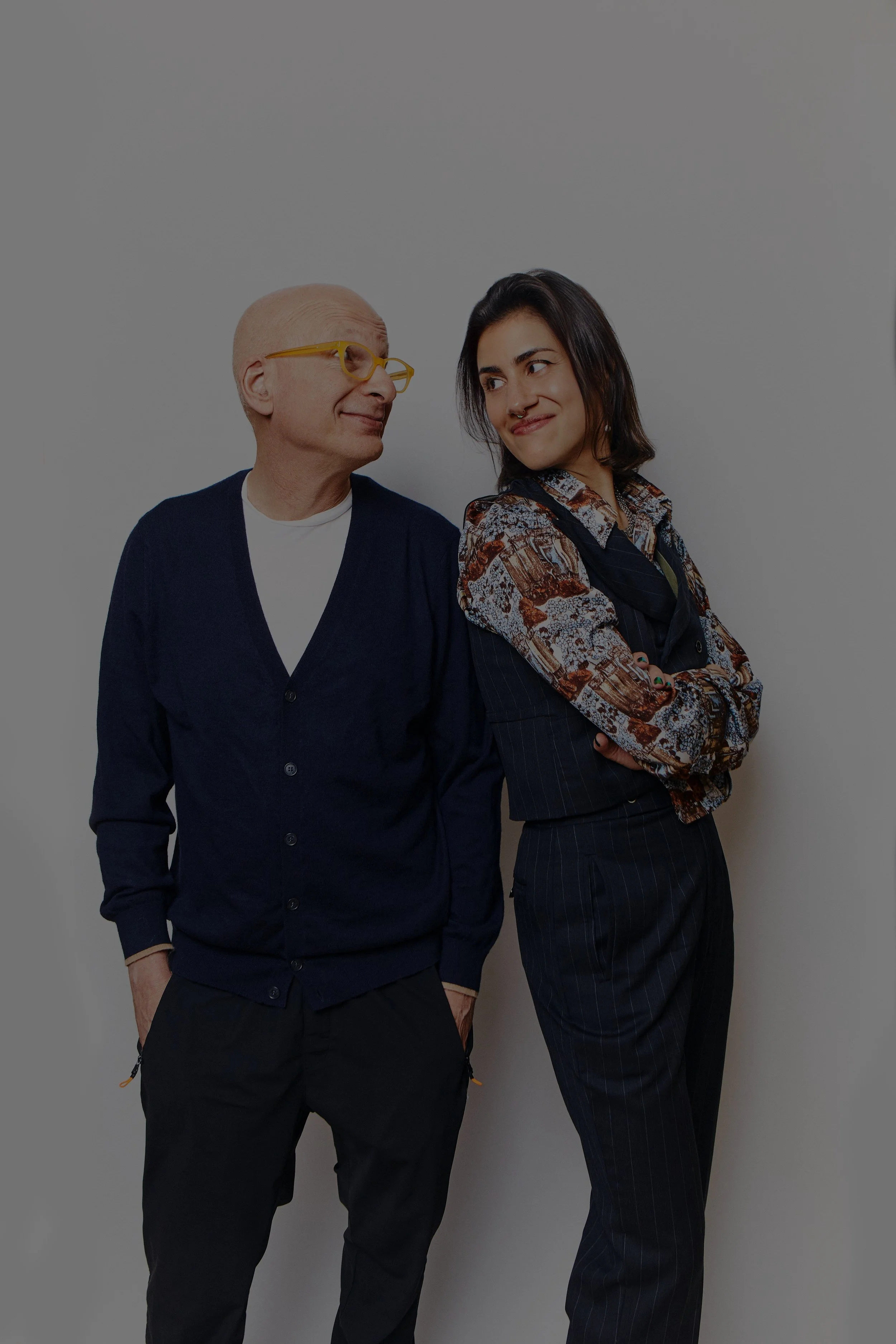 A young woman with dark hair, wearing a patterned button-up shirt and dark pinstripe pants, standing with her arms crossed, smiling at an older man with glasses, who is wearing a dark cardigan over a white shirt, and black pants, smiling back at her in front of a plain gray wall.
