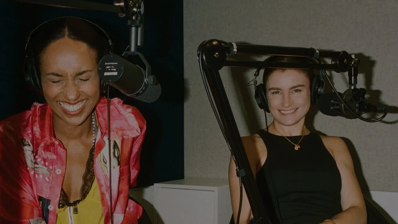 Two women in a recording studio, both wearing headphones and speaking into microphones, one smiling with eyes closed and the other smiling with eyes open.