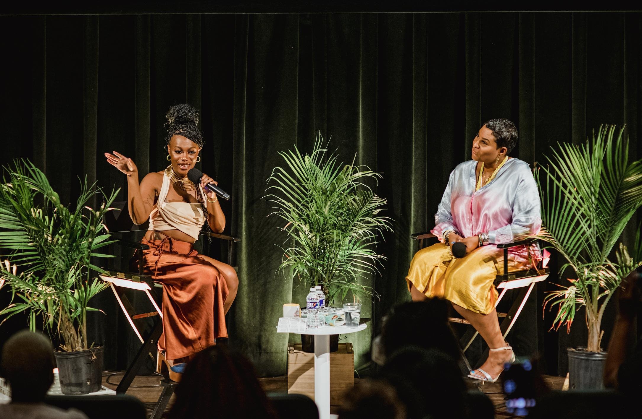 Two women sitting on stage surrounded by green plants, engaged in conversation during a panel discussion. One woman is speaking into a microphone, while the other listens. Audience members are visible in the foreground.