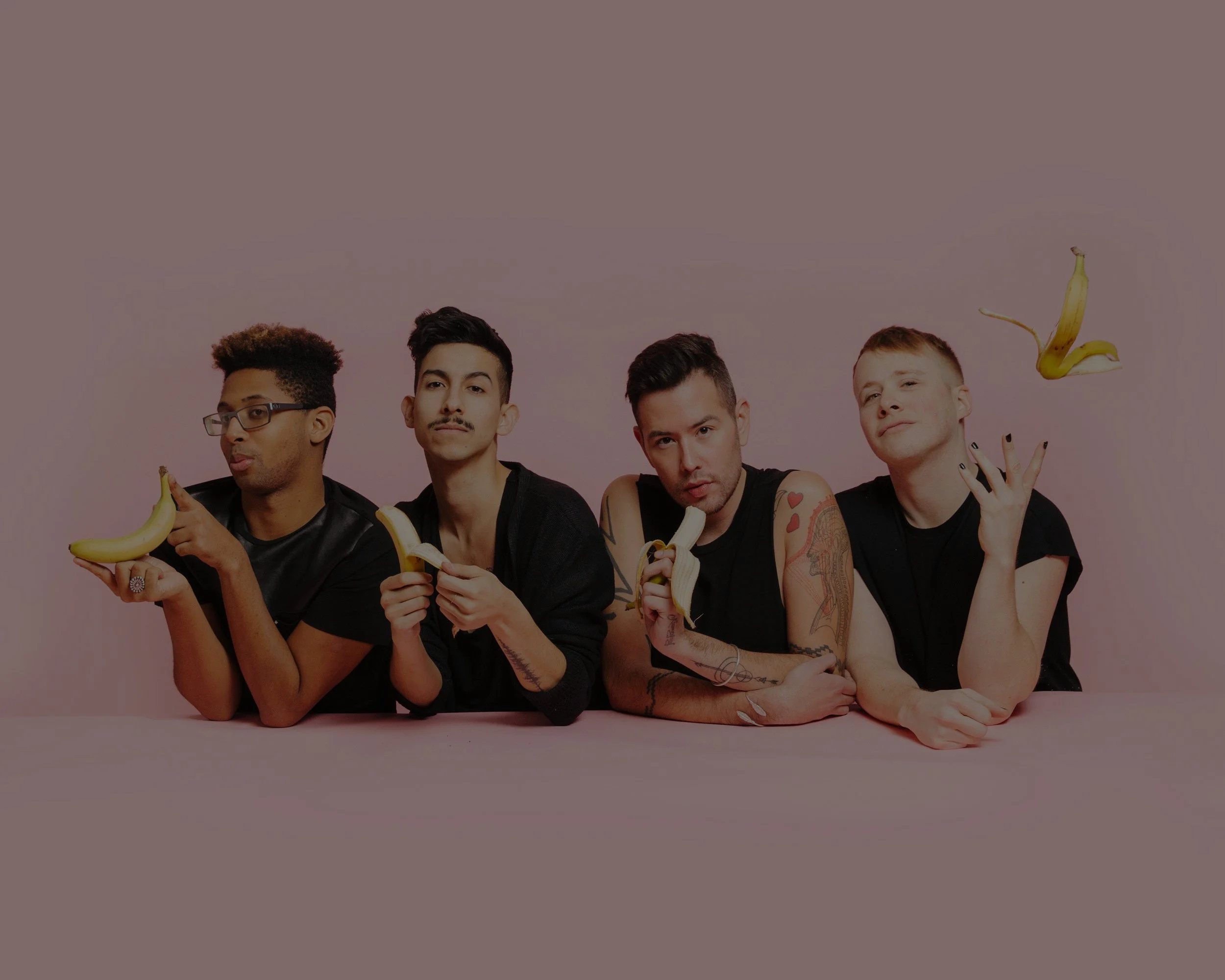 Group of four diverse queers sitting at a table, holding bananas, with a pink background.