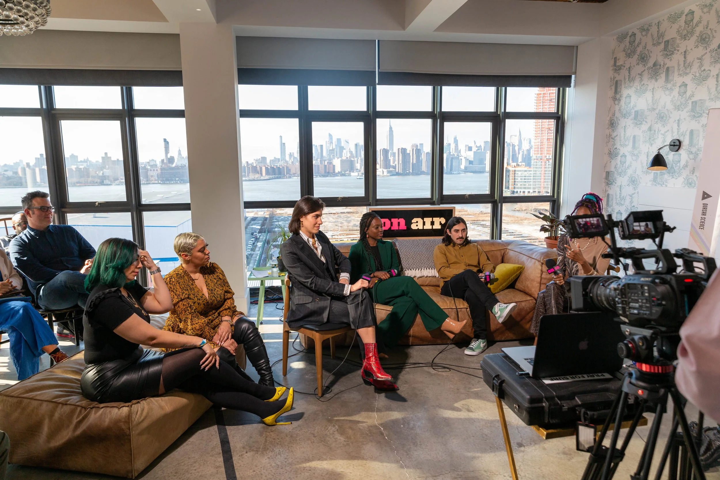 A diverse group of people attending a panel or interview in a room with large windows overlooking a city skyline and water. Some are seated on a sofa, others on chairs, and a camera is recording the event.