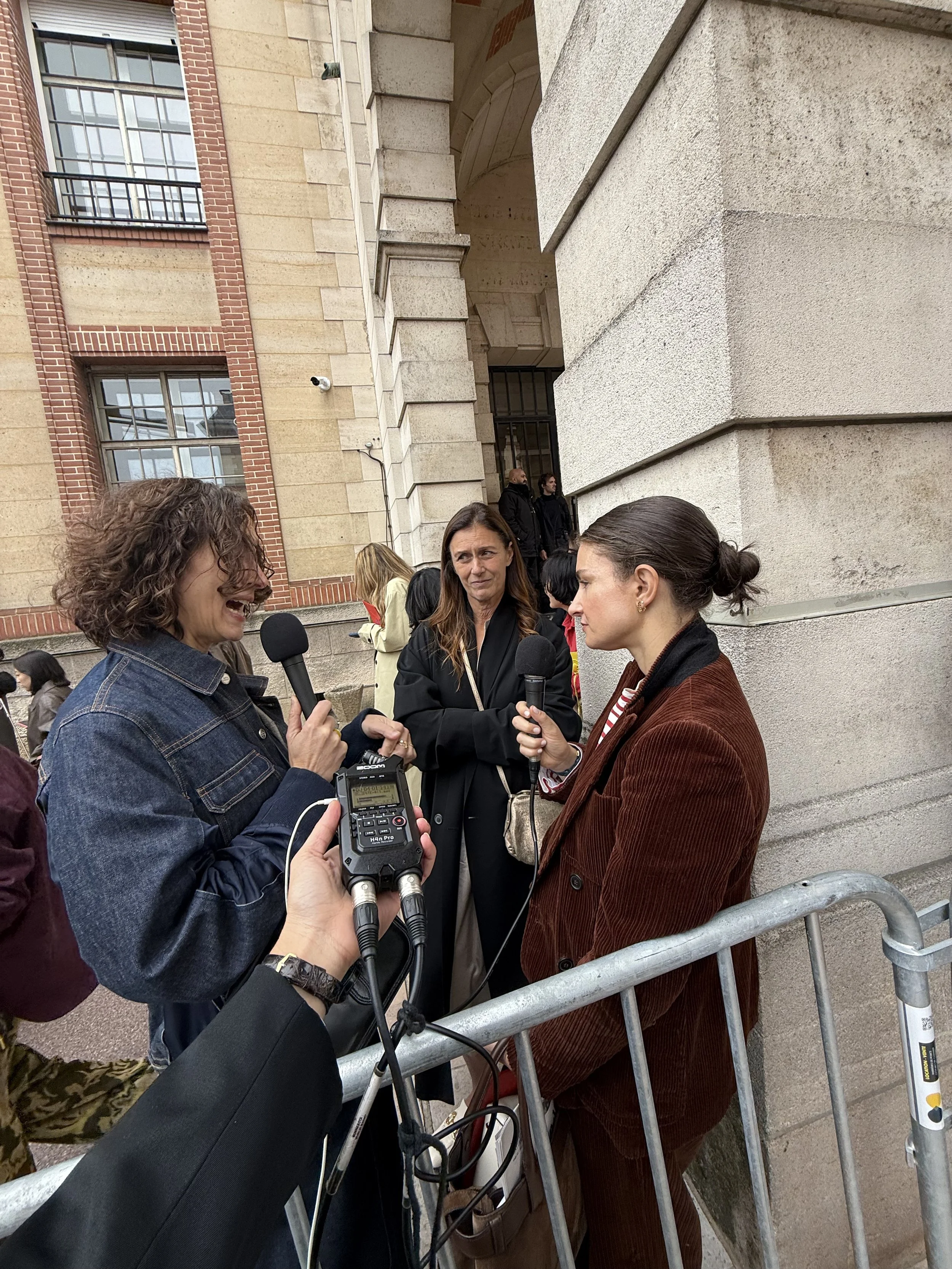 Three women engaged in a conversation or interview outside a building with a brick and stone facade, with other people and security barriers in the background.