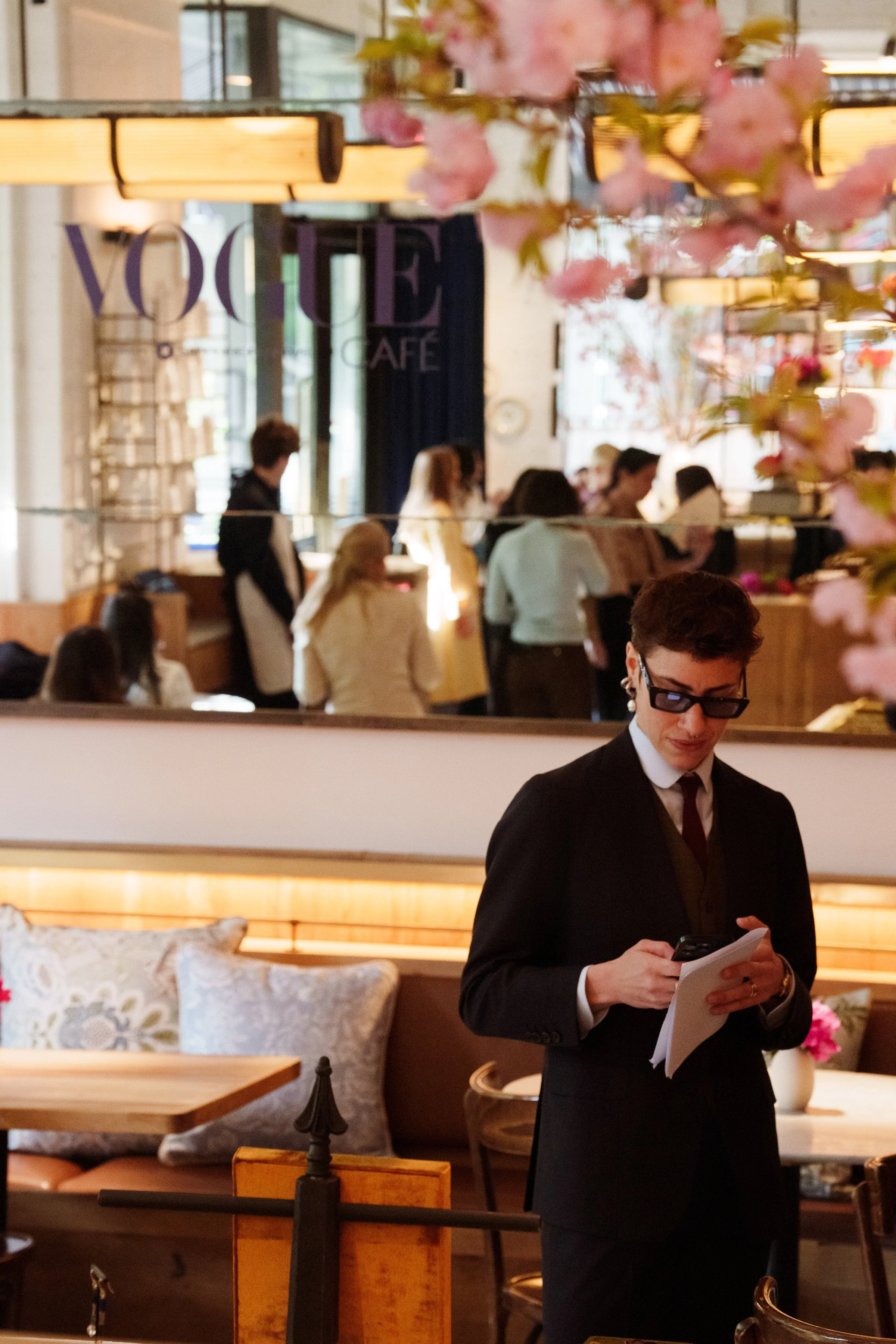 A person dressed in black, wearing glasses and a tie, holding a smartphone and papers, standing inside a cafe with pink flowers and patrons in the background.
