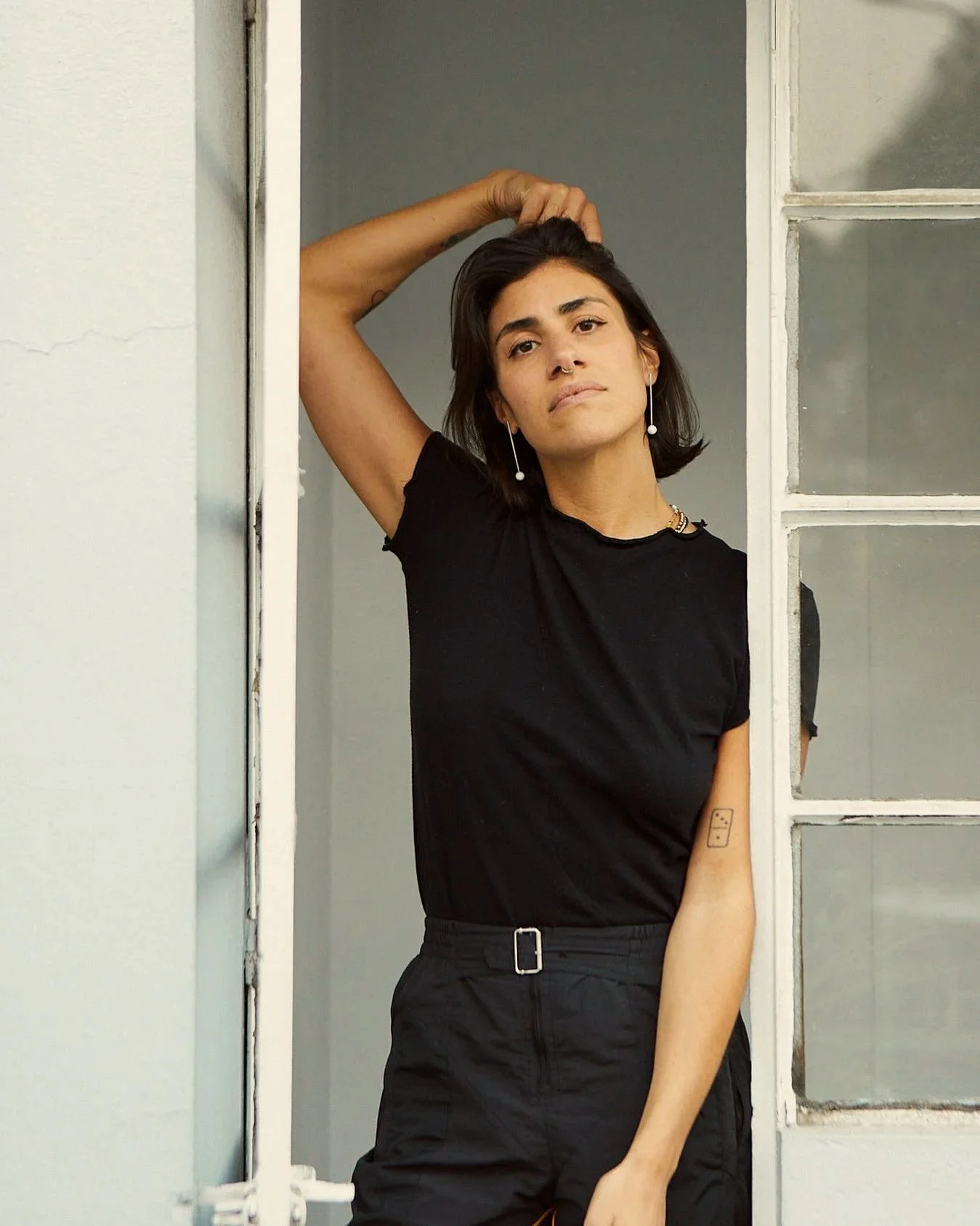 A young woman with short dark hair, wearing a black t-shirt and black pants, standing inside a room with a window. She is holding her hair with her right hand and looking at the camera with a neutral expression.