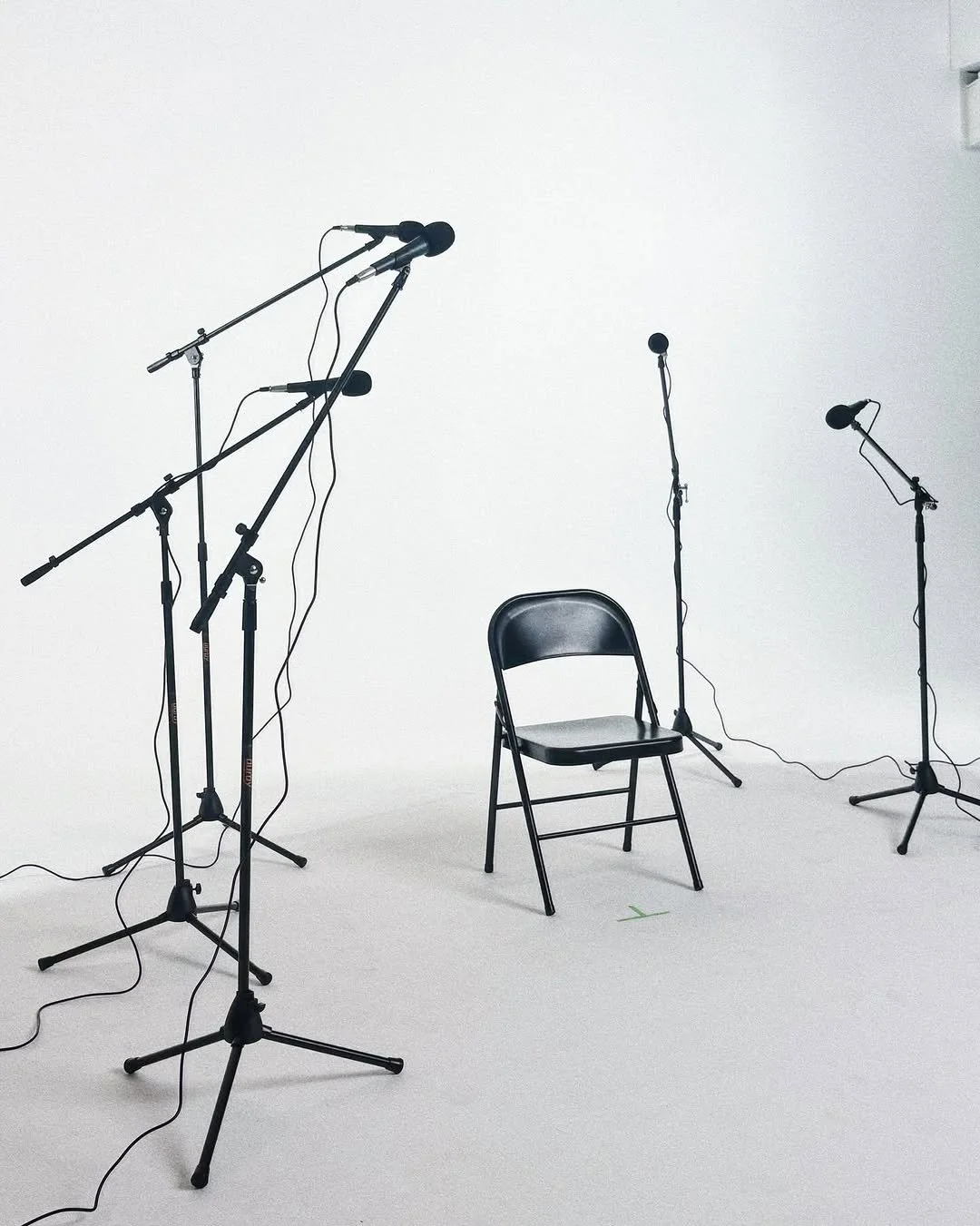 Empty black chair surrounded by multiple microphone stands in a white studio