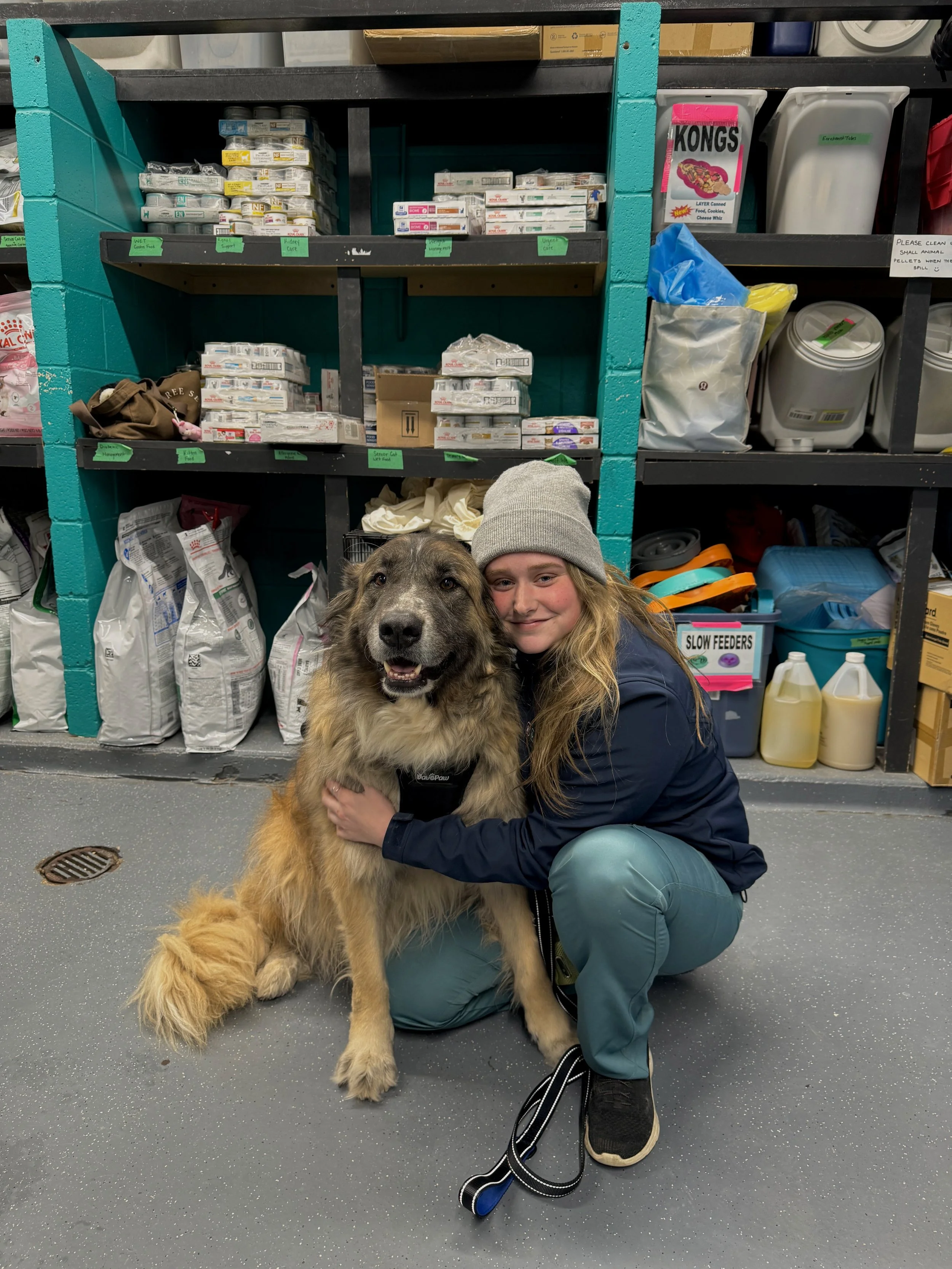 A happy client after graduating from Karen Pryor training programs. A woman and a large fluffy dog sitting on the floor of a pet store aisle, with shelves full of pet food and supplies behind them. Book now in Oakville, Hamilton, Burlington, Milton